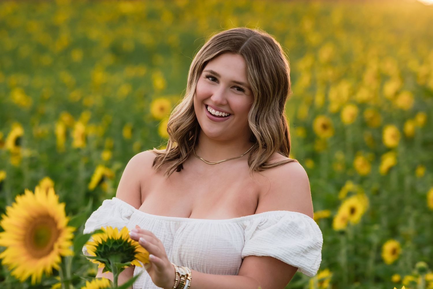 A person poses in a white off-shoulder dress among vibrant yellow sunflowers on a sunny summer day.