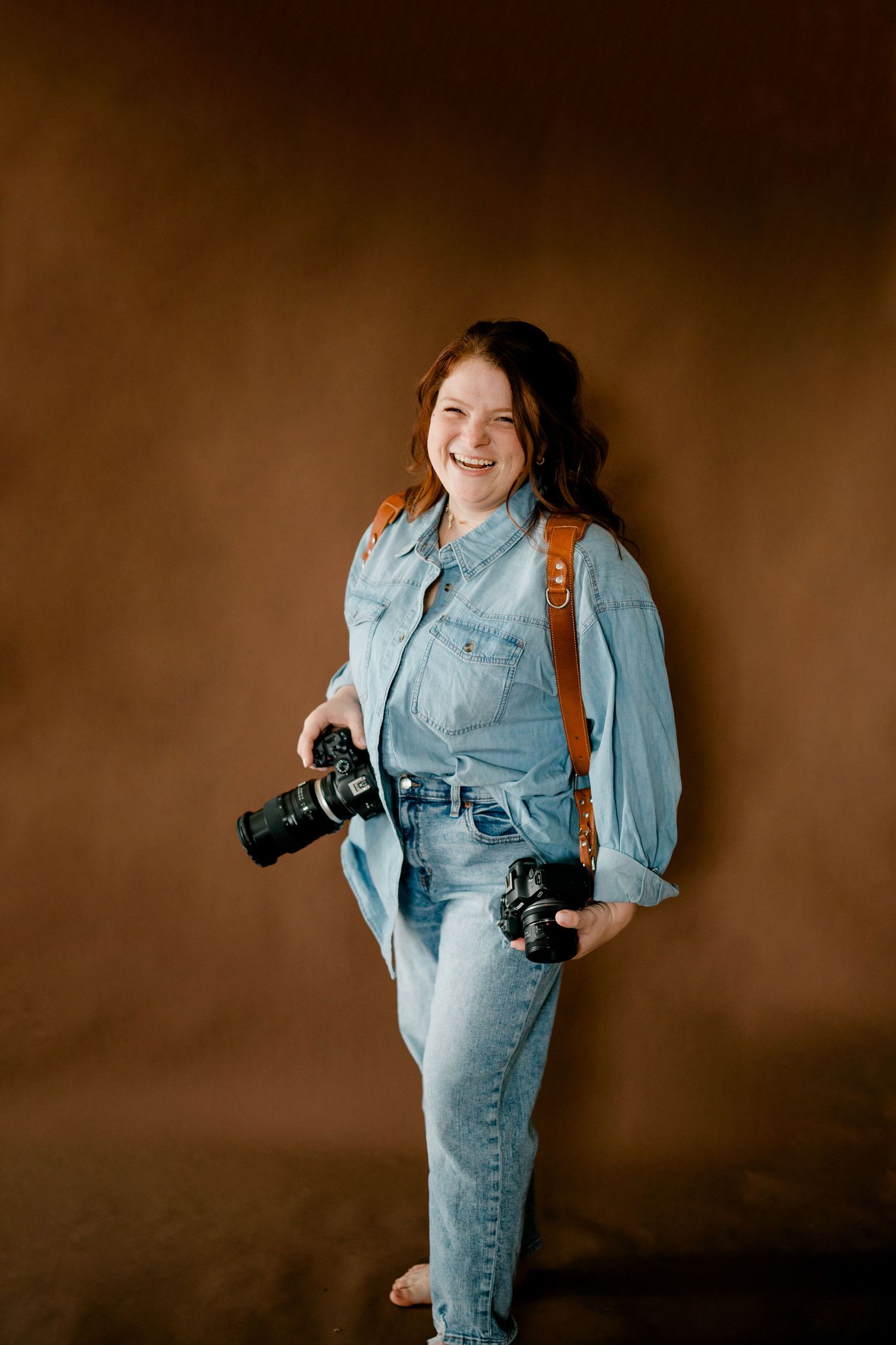 Cheerful female photographer in denim outfit holding two cameras against a warm brown backdrop.