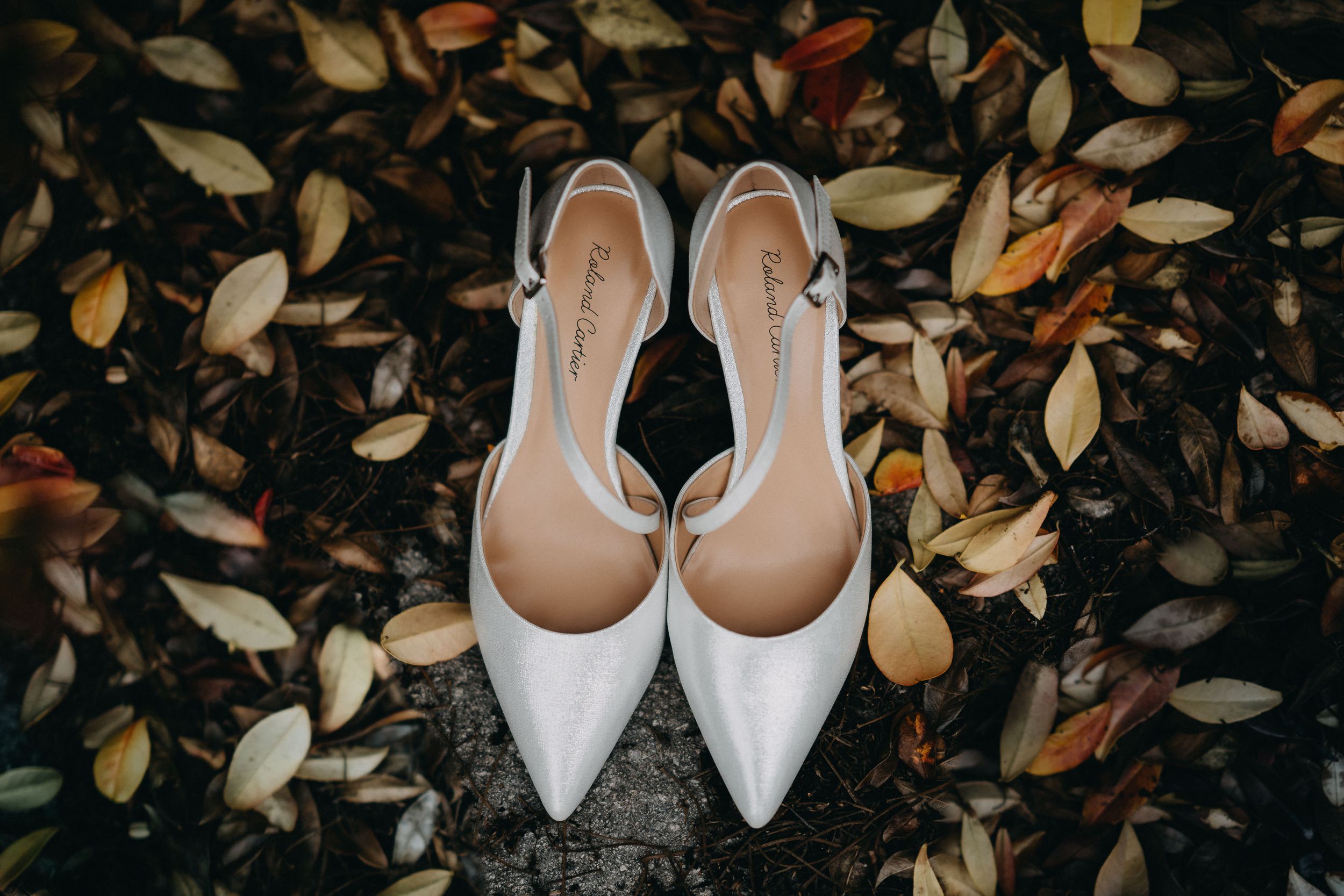 White pointed-toe bridal shoes placed on autumn leaves.