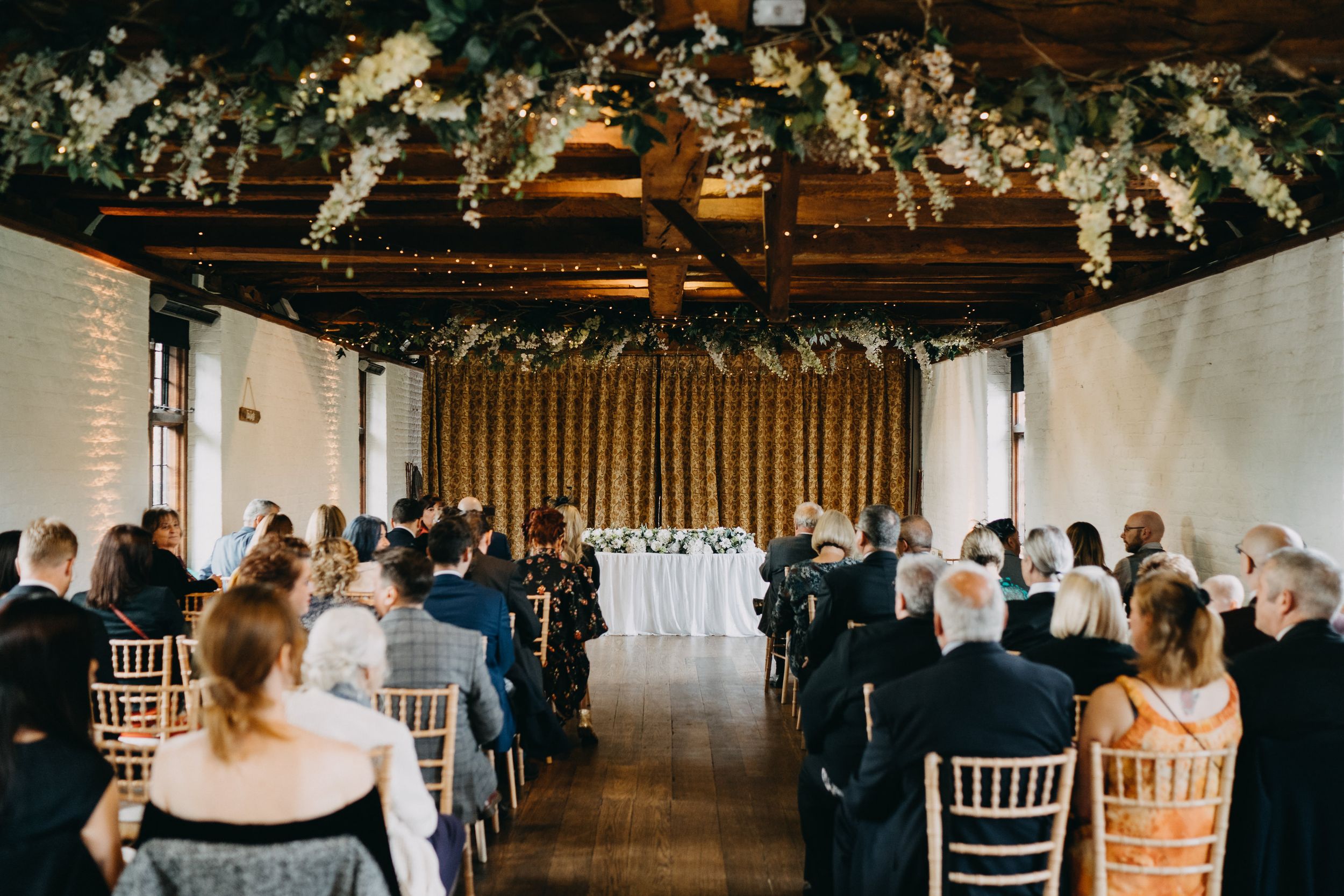Wedding ceremony at Tudor Barn Eltham venue with floral ceiling decorations and rows of white chairs.