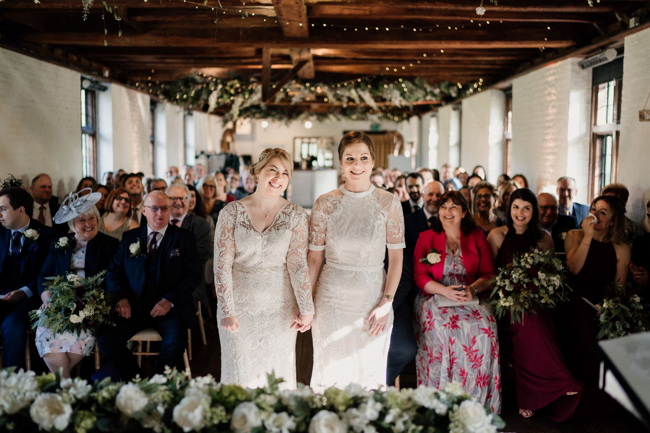 Two people in elegant white lace wedding dresses stand together at the altar during an indoor ceremony.