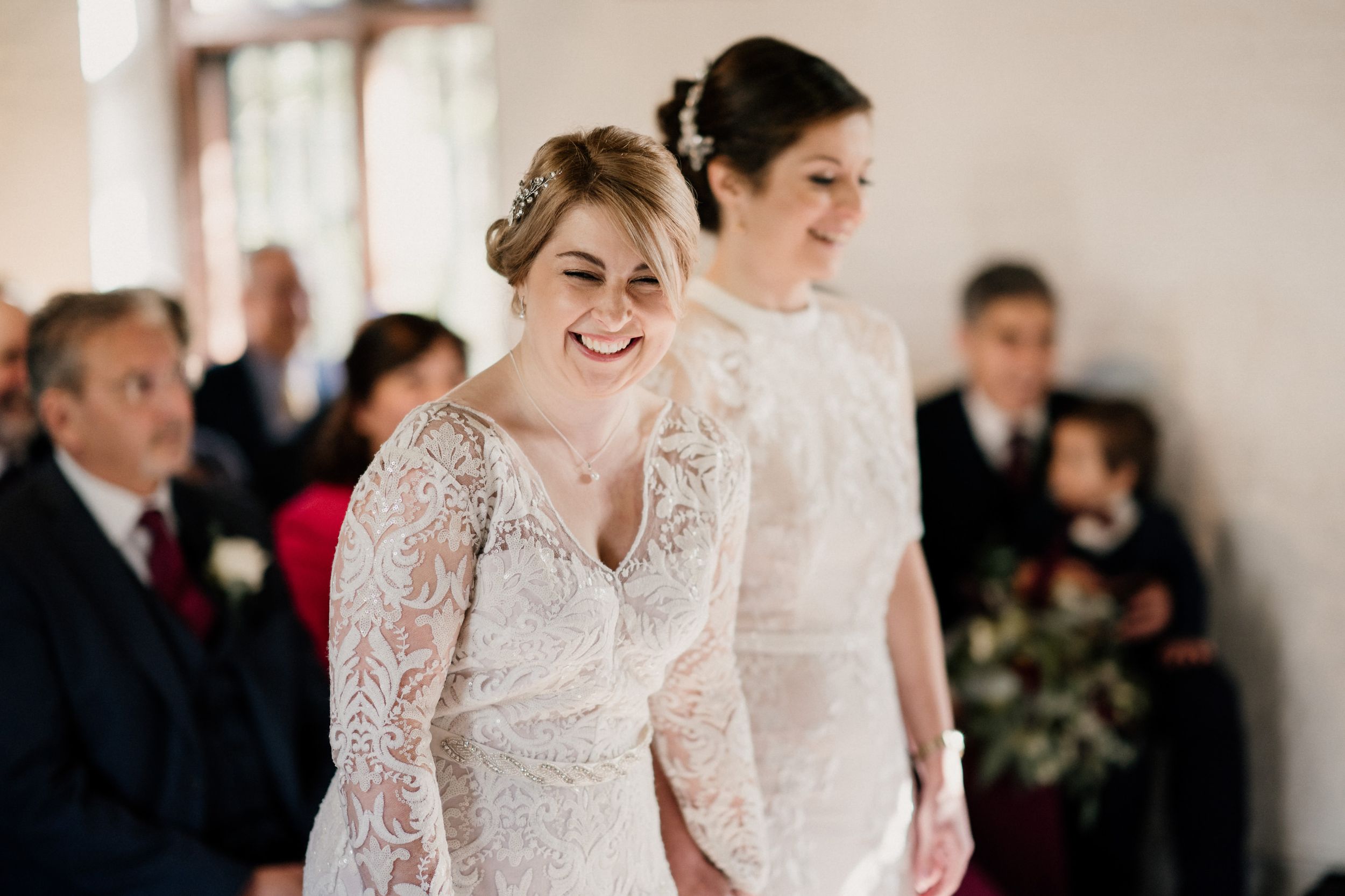 Two people wearing long-sleeved lace wedding gowns share a joyful moment during the ceremony.