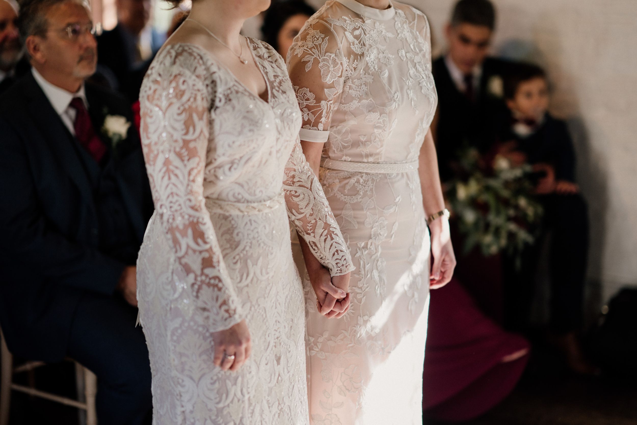 Close-up of two people holding hands while wearing matching white lace wedding gowns.