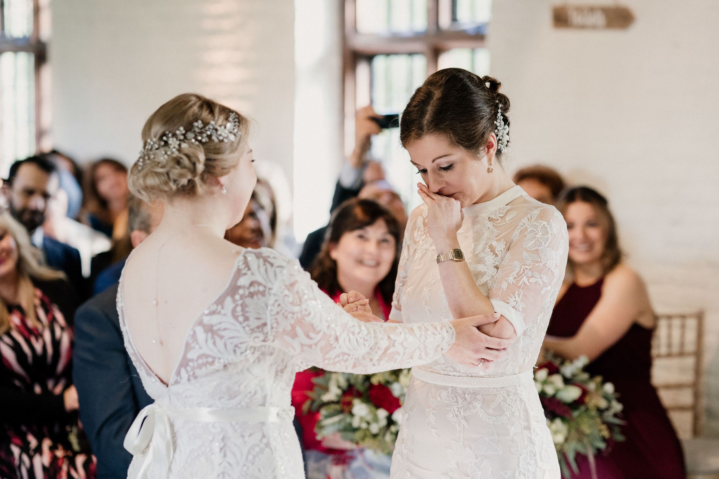 Two brides in white lace wedding dresses share an emotional moment during their ceremony.