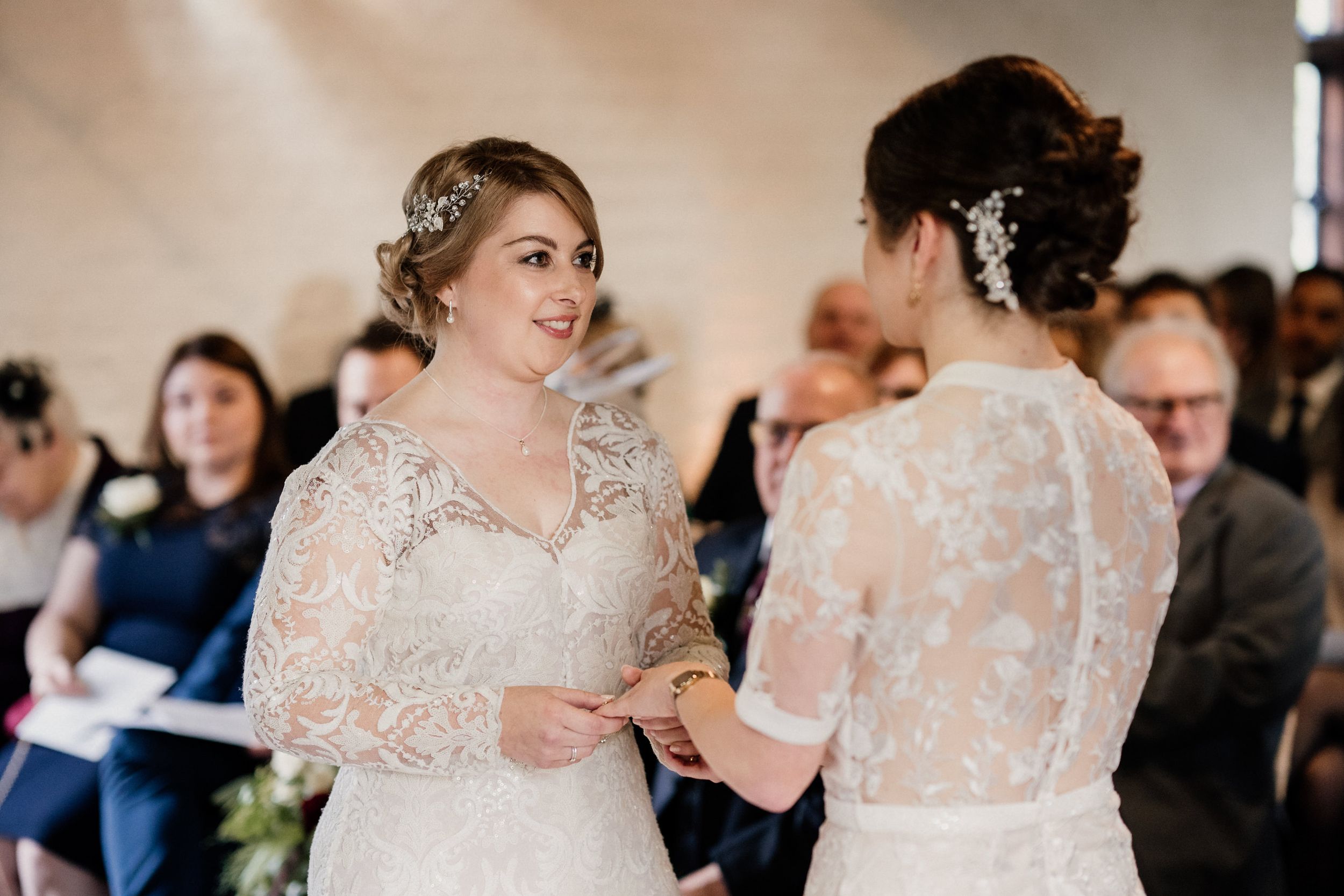 Two brides in delicate lace wedding gowns exchange vows during an indoor ceremony.