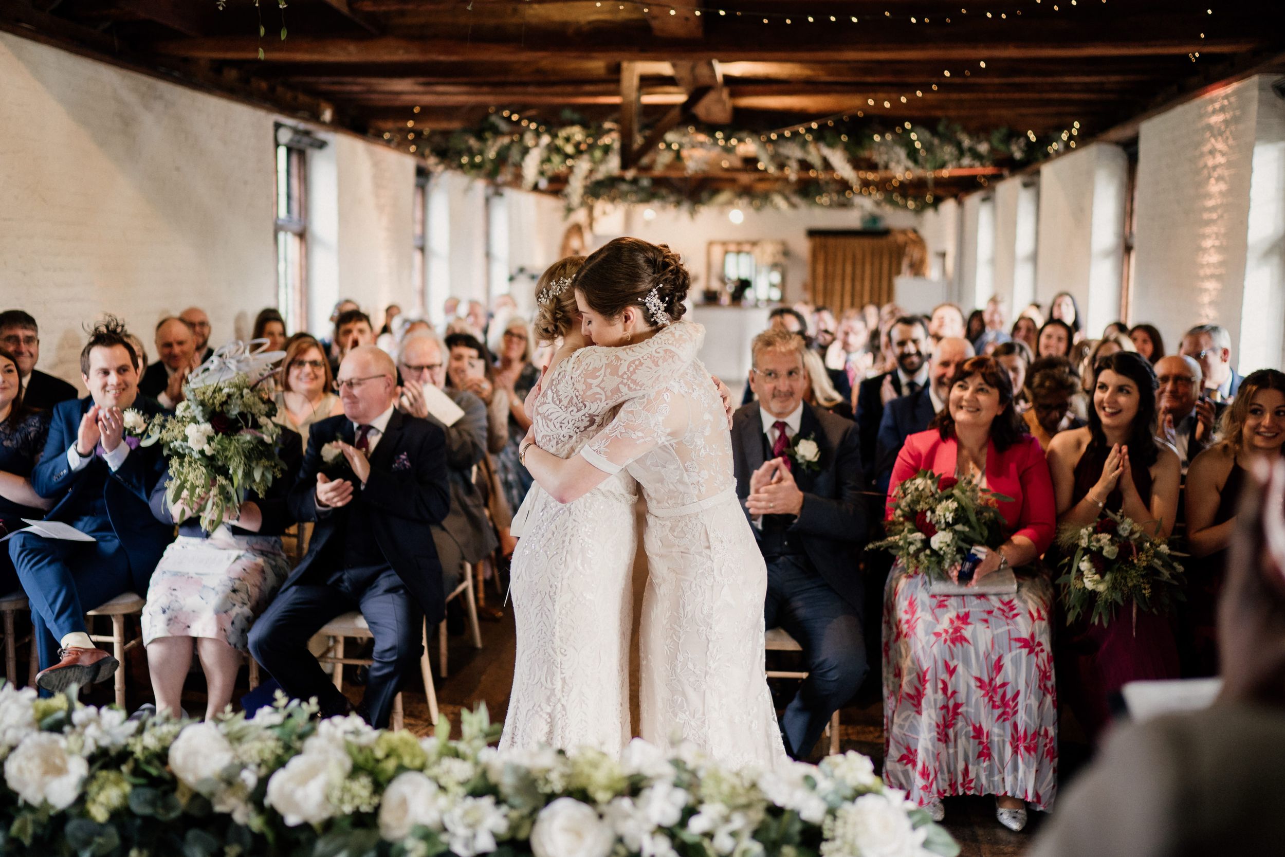 Two brides in white wedding dresses embrace during their ceremony as guests look on.