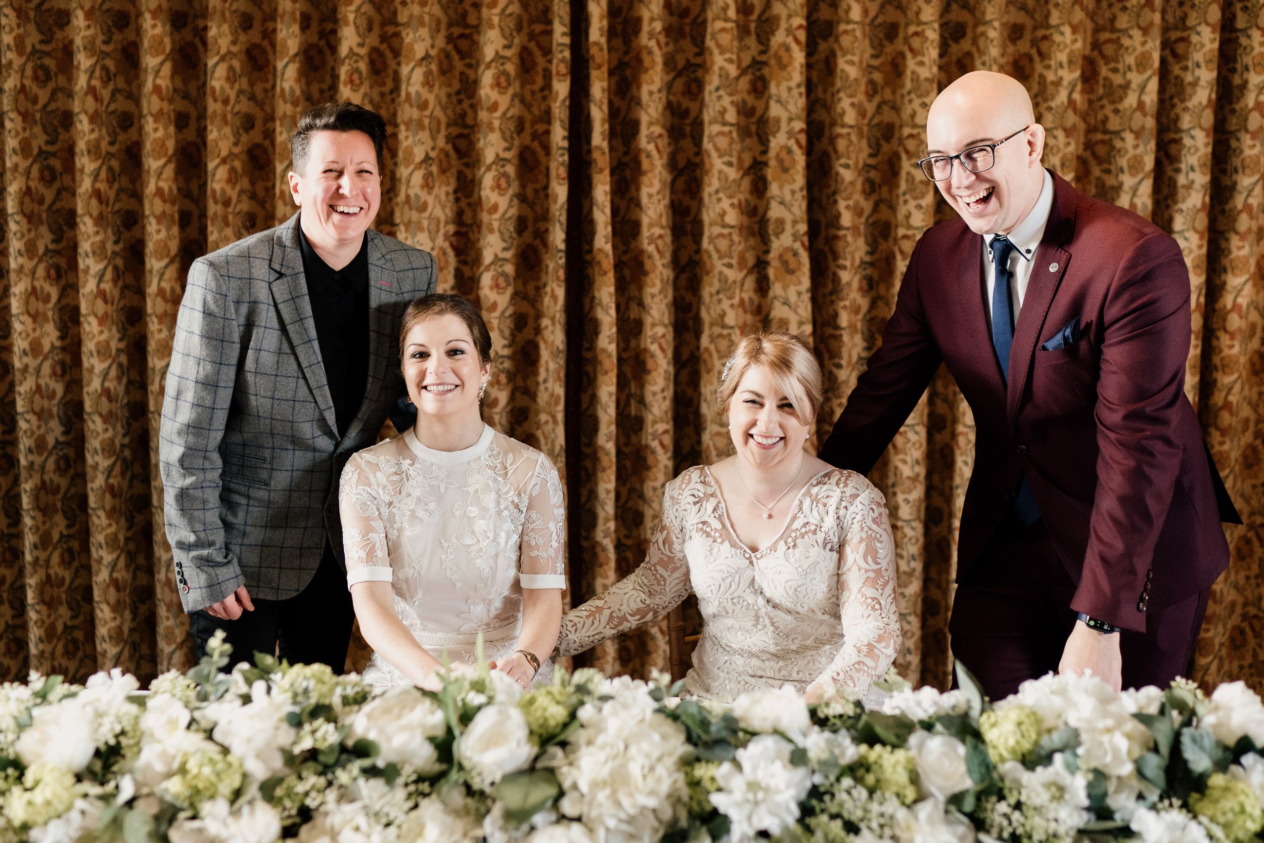 Wedding party poses together at reception table decorated with white floral arrangements.