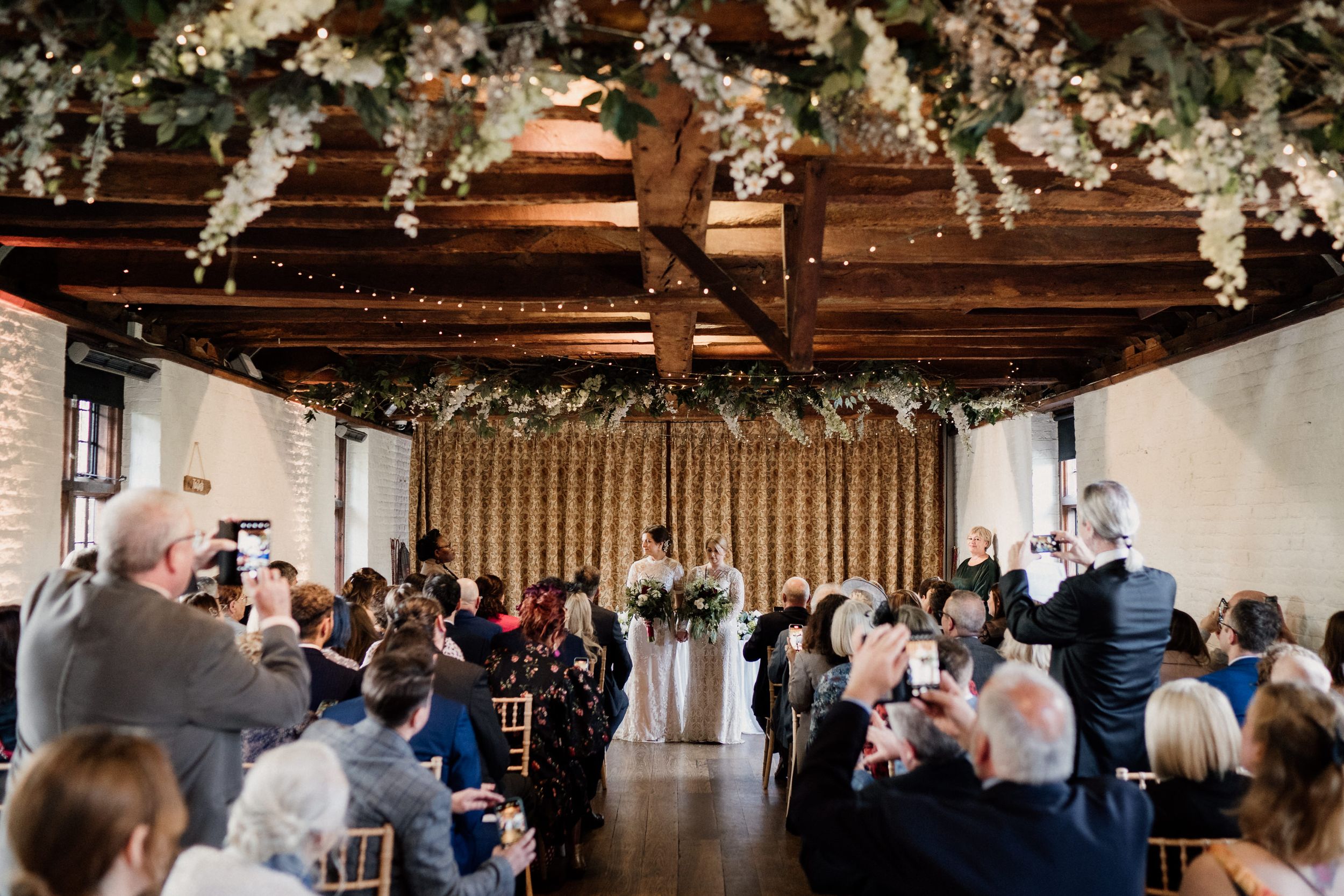 Wedding ceremony at Tudor Barn London in progress inside venue with wooden beams and floral decorations.
