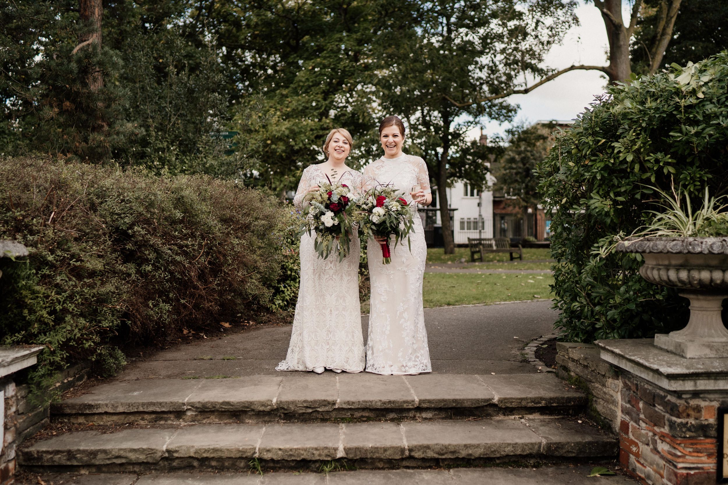 Two brides in white wedding dresses stand together holding bouquets on stone steps outdoors.