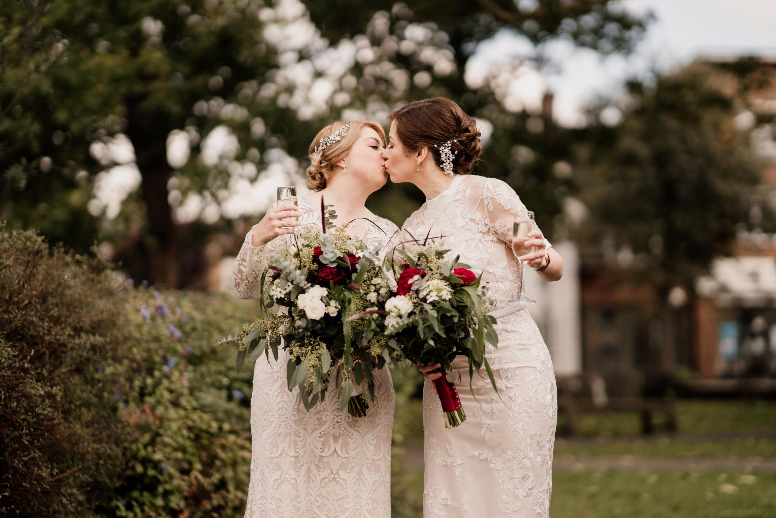 Two brides in white lace dresses share a tender moment while holding floral bouquets in an outdoor garden setting of Tudor Barn Eltham.