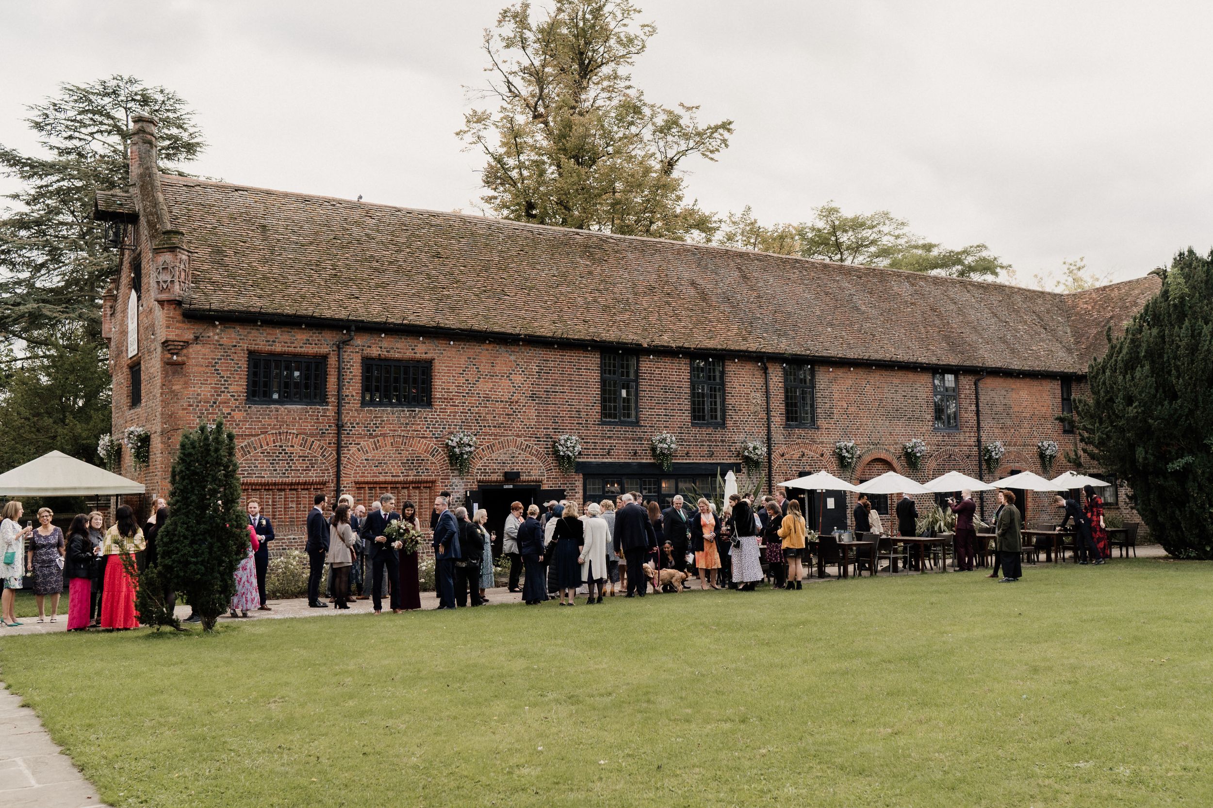 Wedding guests gather outside rustic Tudor Barn with white umbrellas on lawn.
