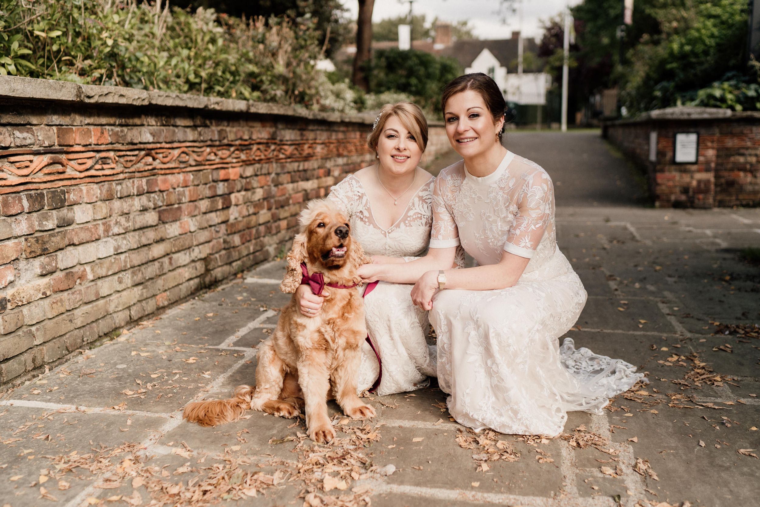 A happy dog sits between two people in matching white lace dresses on a stone path.