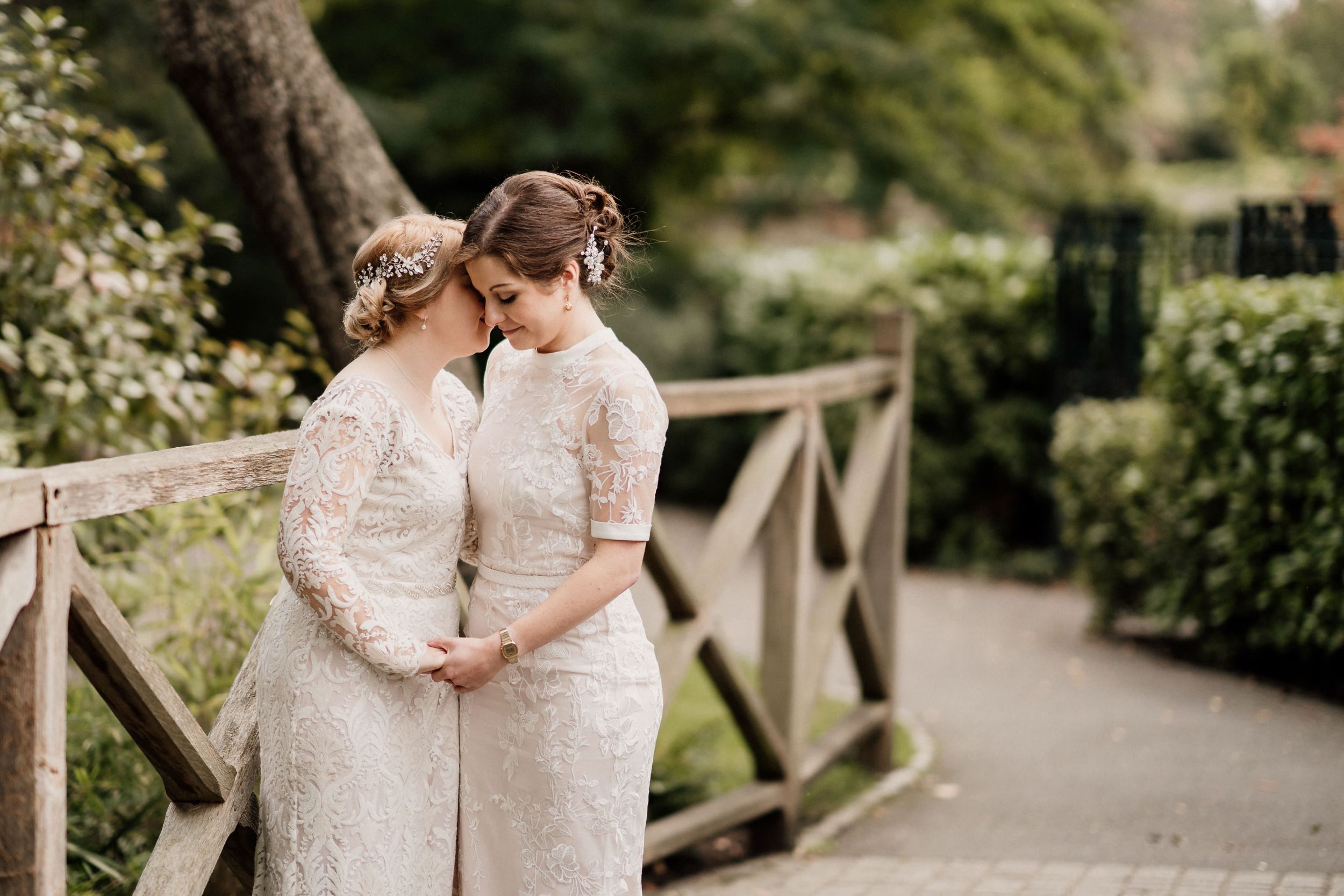 Two people in white lace dresses share an intimate moment on a garden bridge.