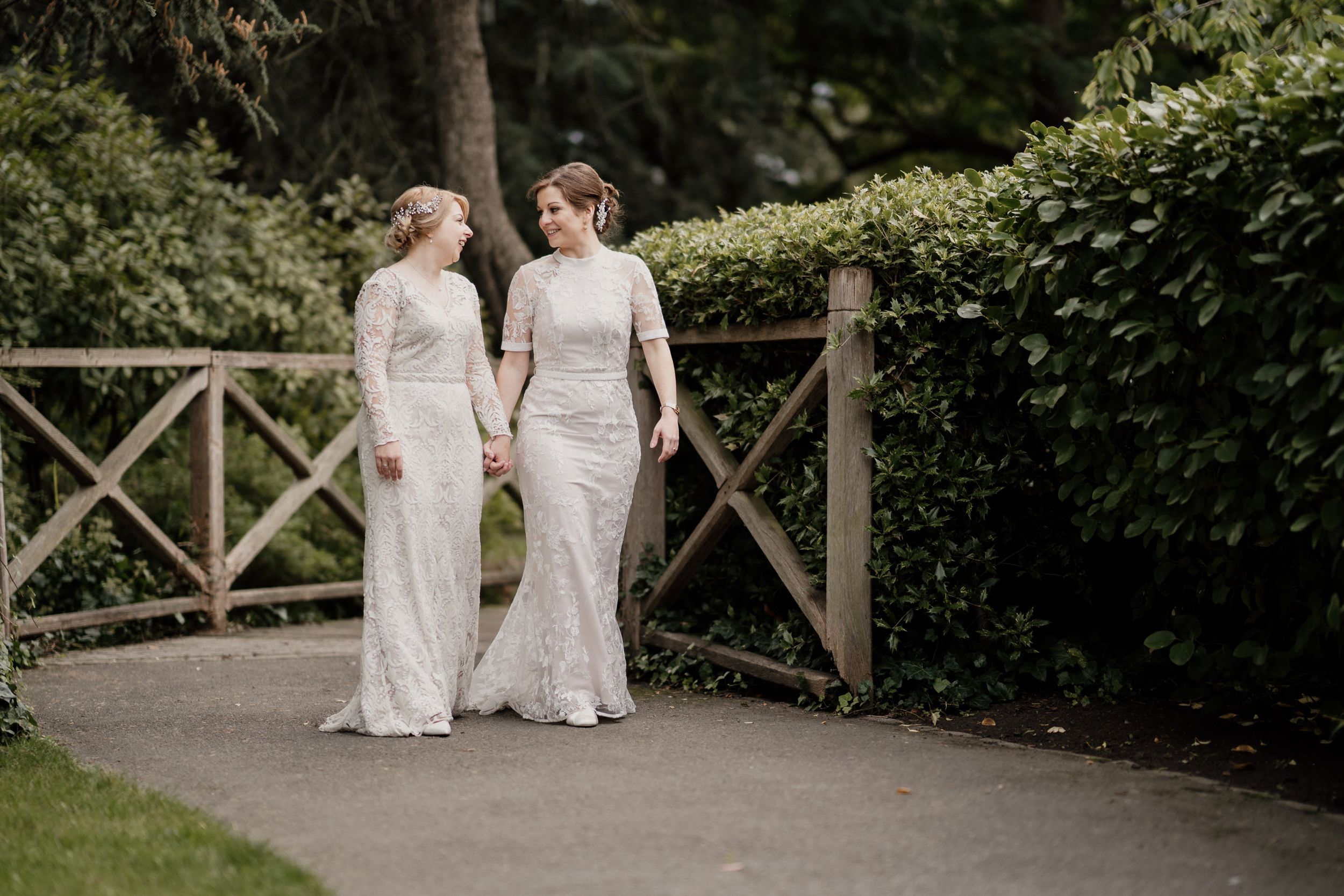 Two people in white lace dresses walk hand in hand along a garden path with wooden fencing.