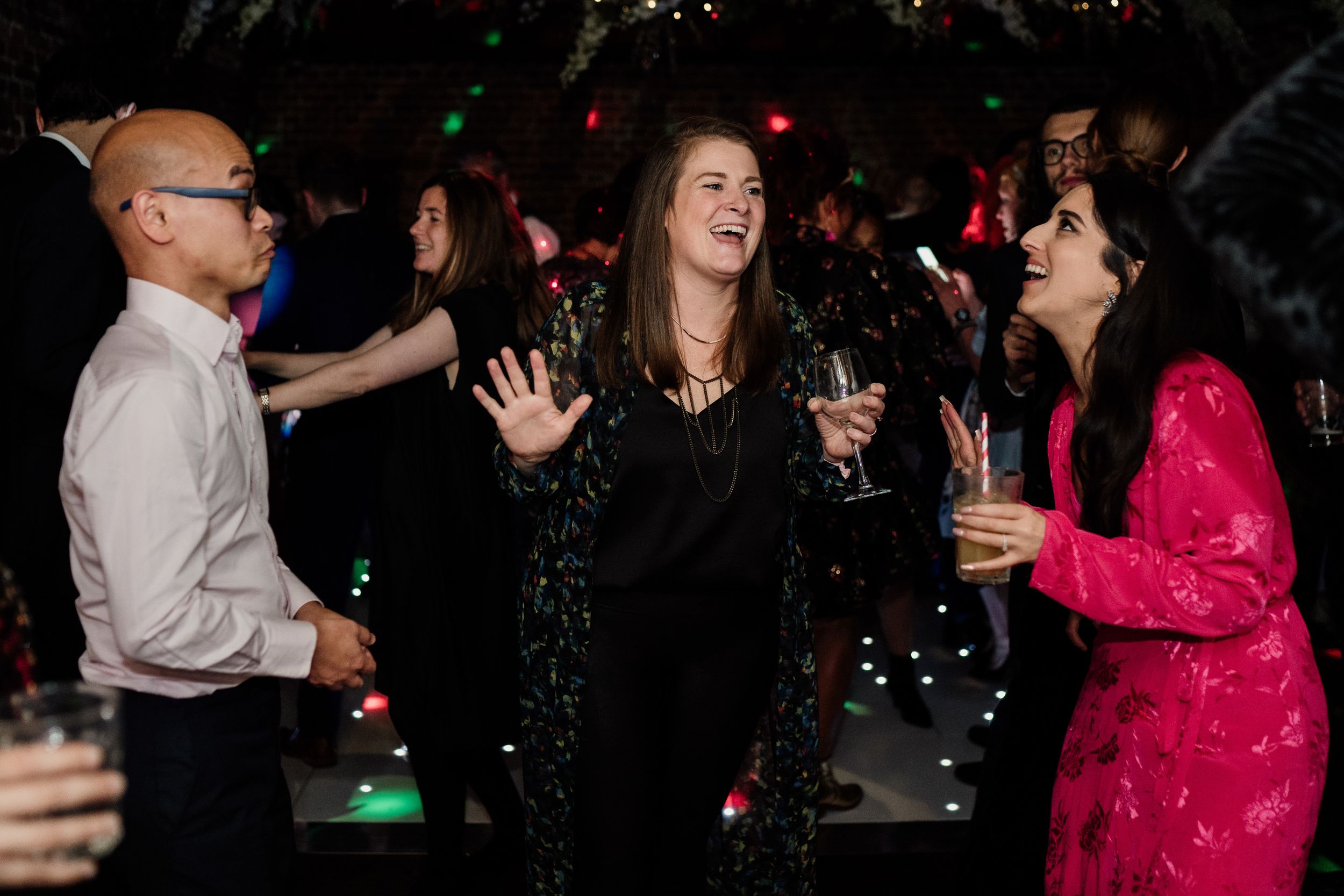 Party guests laughing and dancing at reception with colorful lighting.