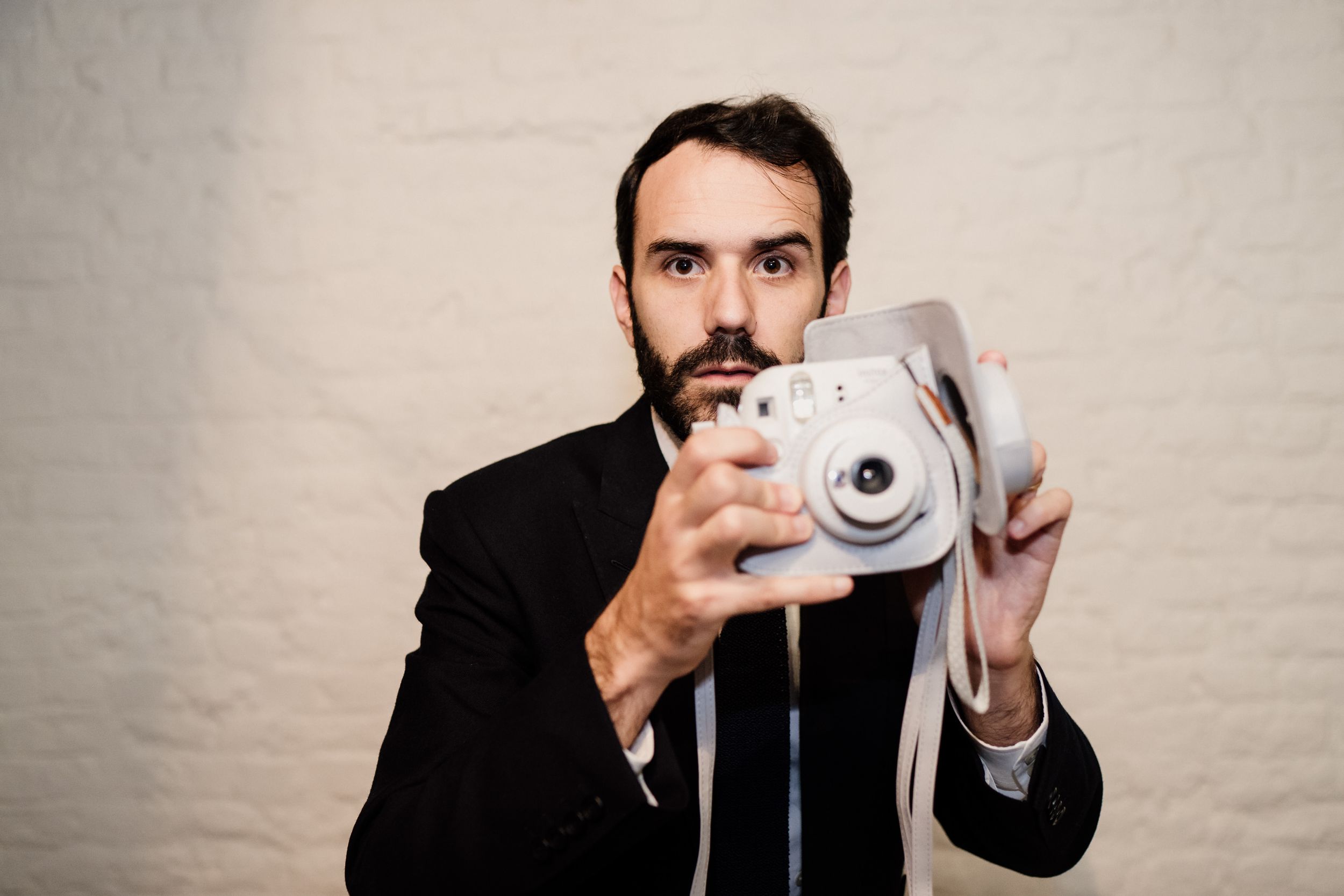 Professional photographer in black suit holding instant camera against white wall.