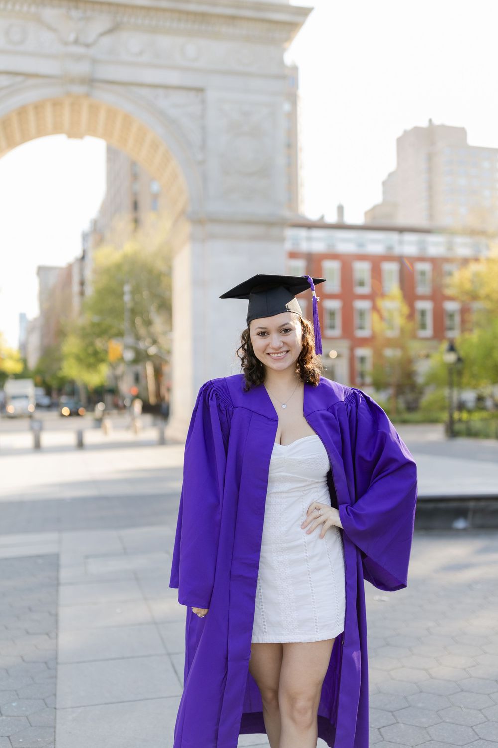 Graduate in purple commencement robe poses in front of historic arch.