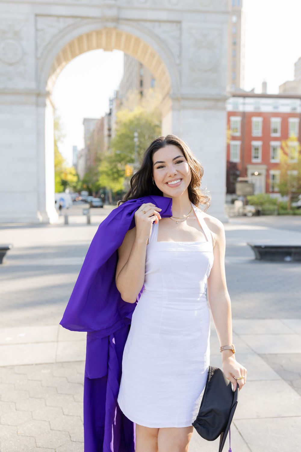 Graduate in white dress holds purple graduation gown over shoulder near arch.