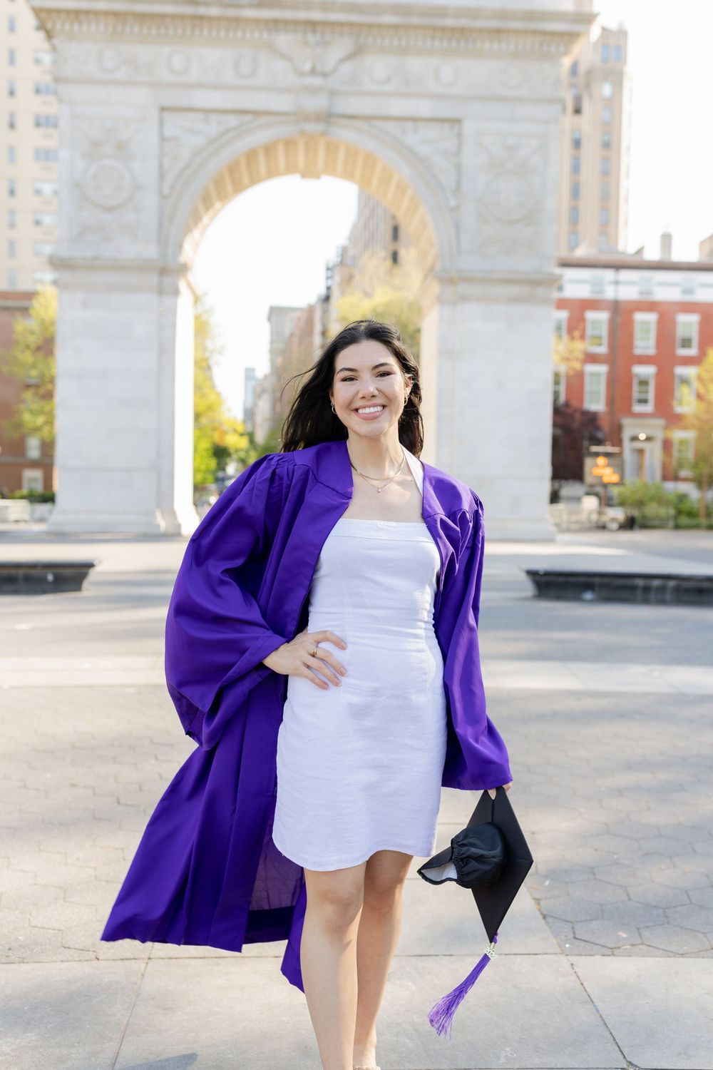 Graduate in white dress and purple NYU graduation gown poses in front of Washington Square Arch.