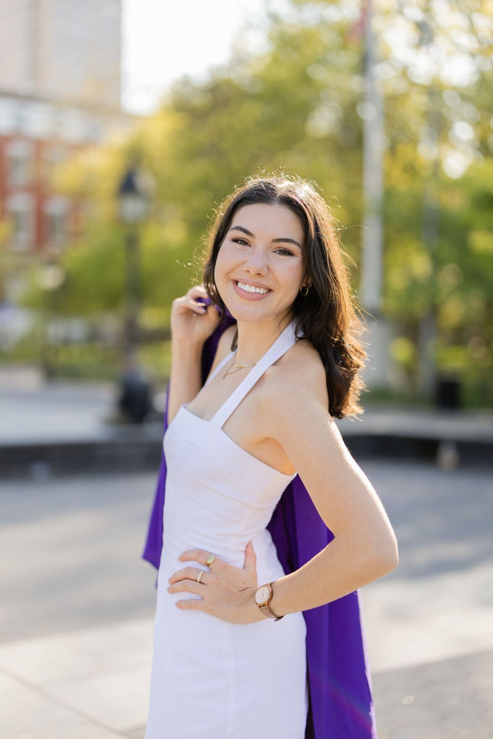 Graduate in white dress poses with purple graduation gown in Washington Square Park.