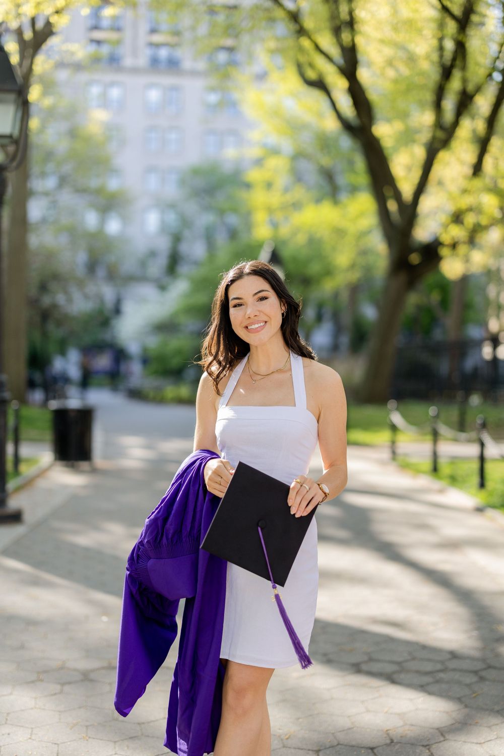 Graduate holds diploma while wearing white dress and purple gown in sunny park setting.