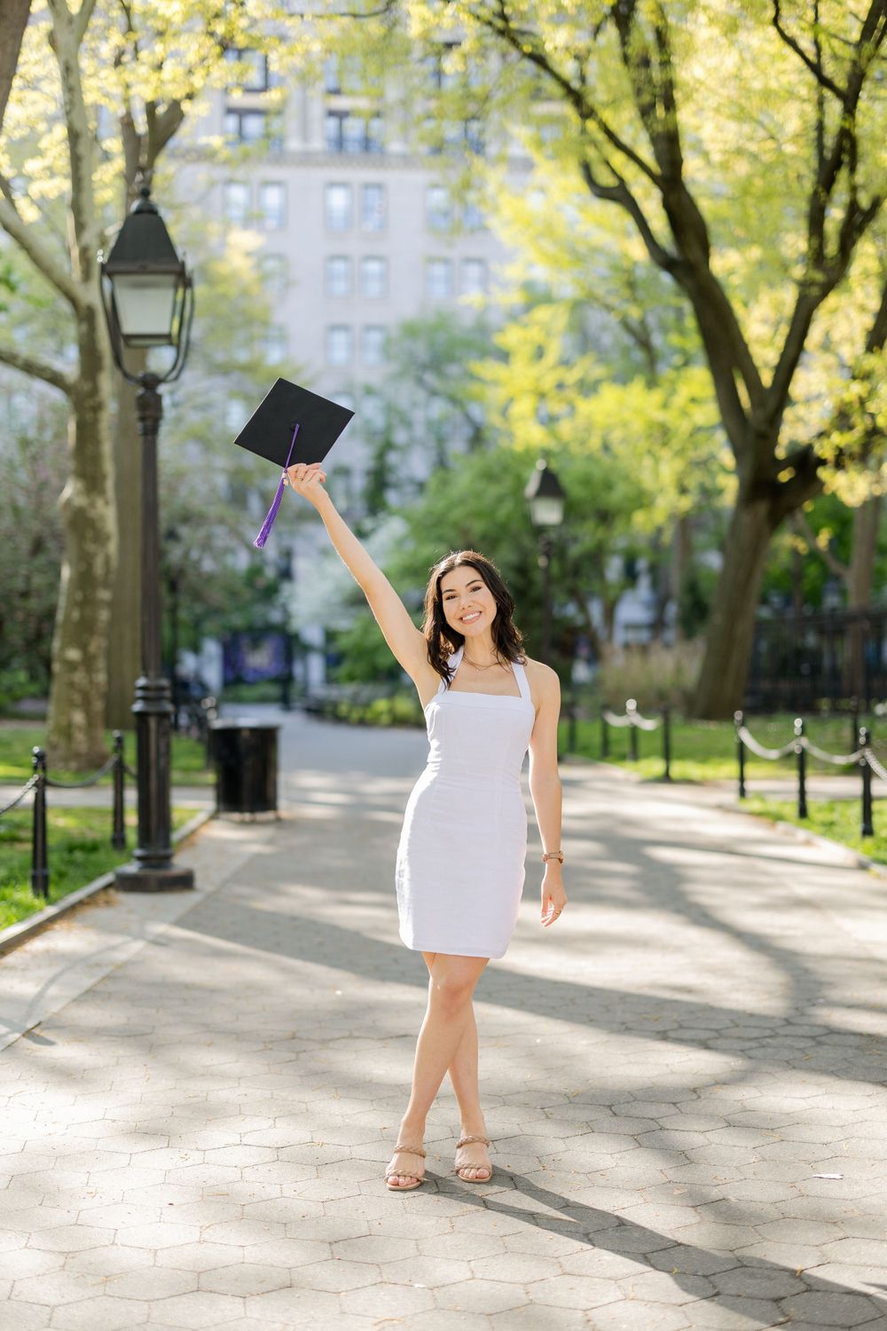 Graduate celebrates by raising graduation cap on tree-lined pathway.