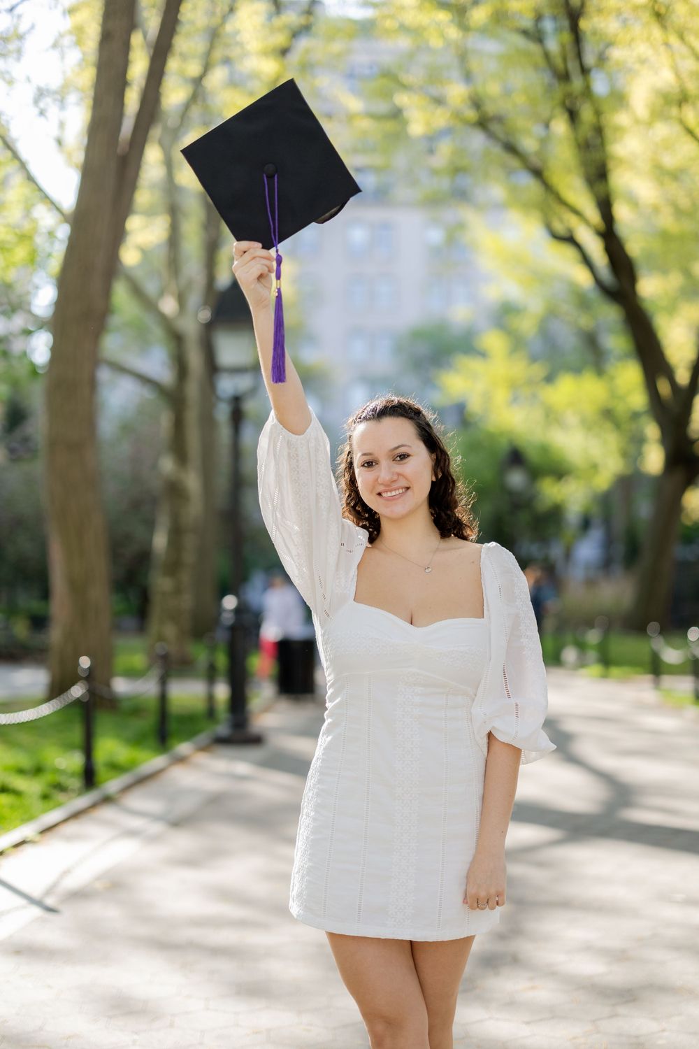 Graduate raises graduation cap while wearing white dress in Washington Square Park.