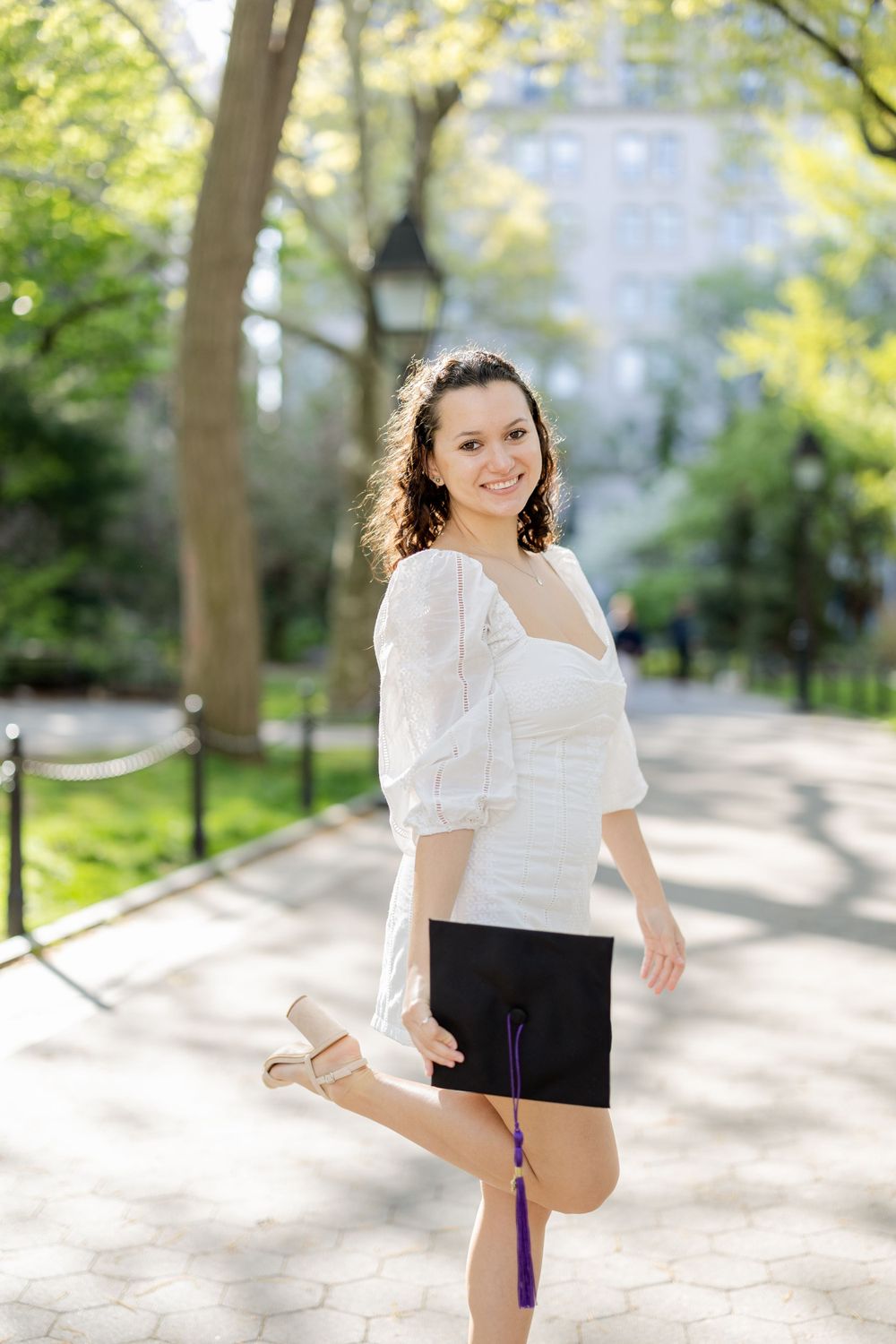 Graduate poses playfully with diploma in park setting wearing white dress.