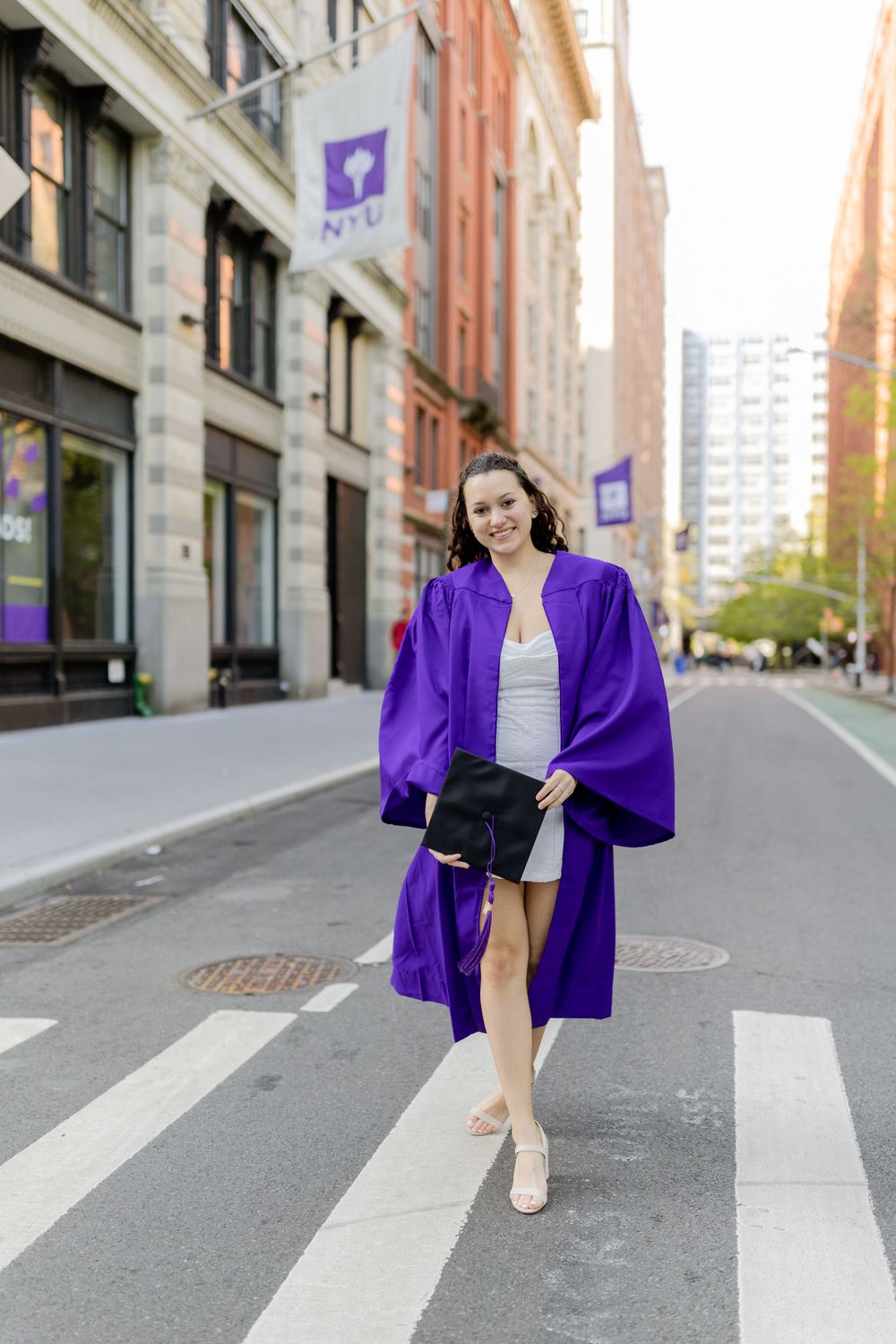 Graduate walks across NYU campus crosswalk in purple graduation regalia.