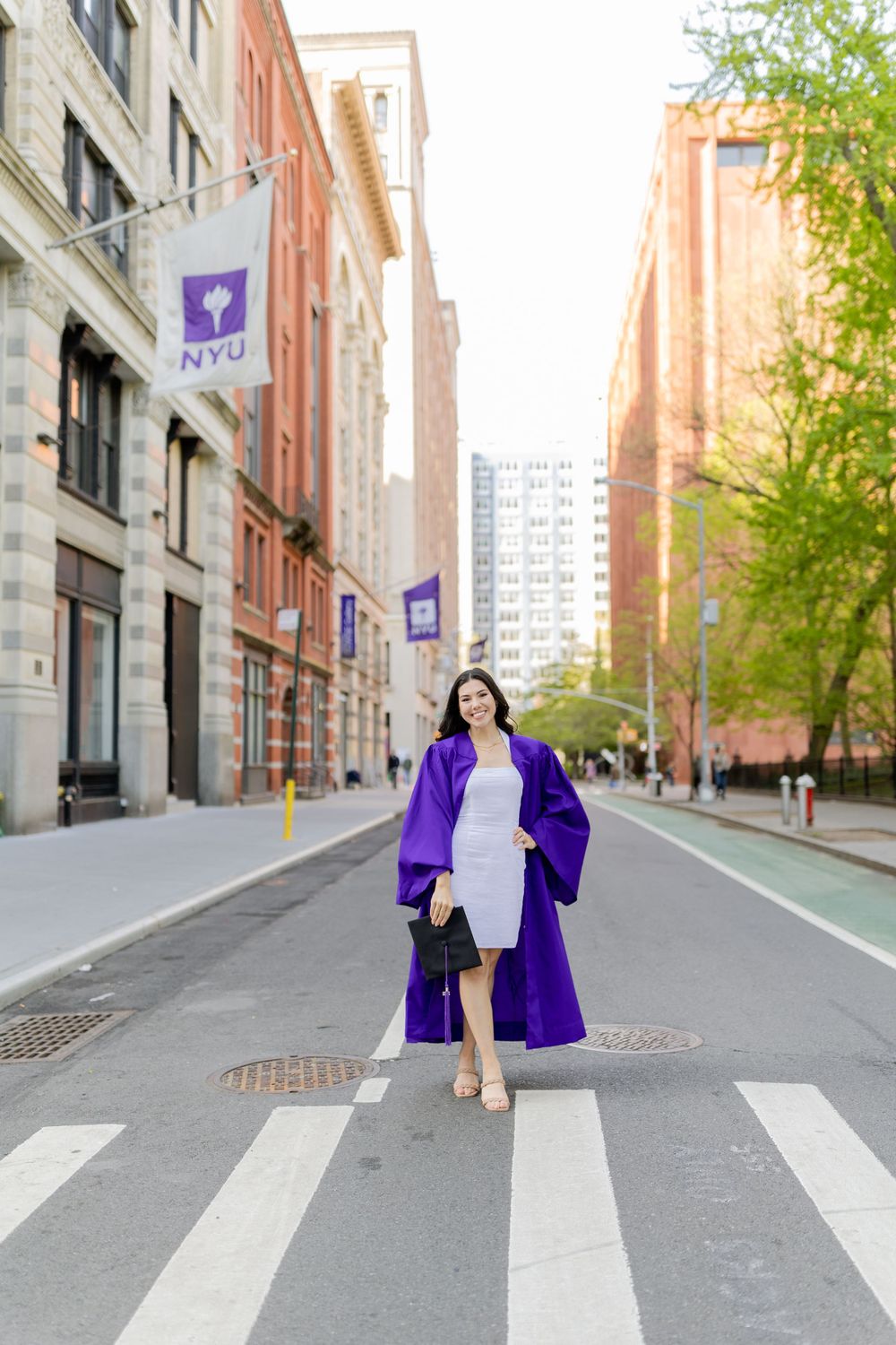 NYU graduate in purple commencement robe crosses street with NYU banners visible.