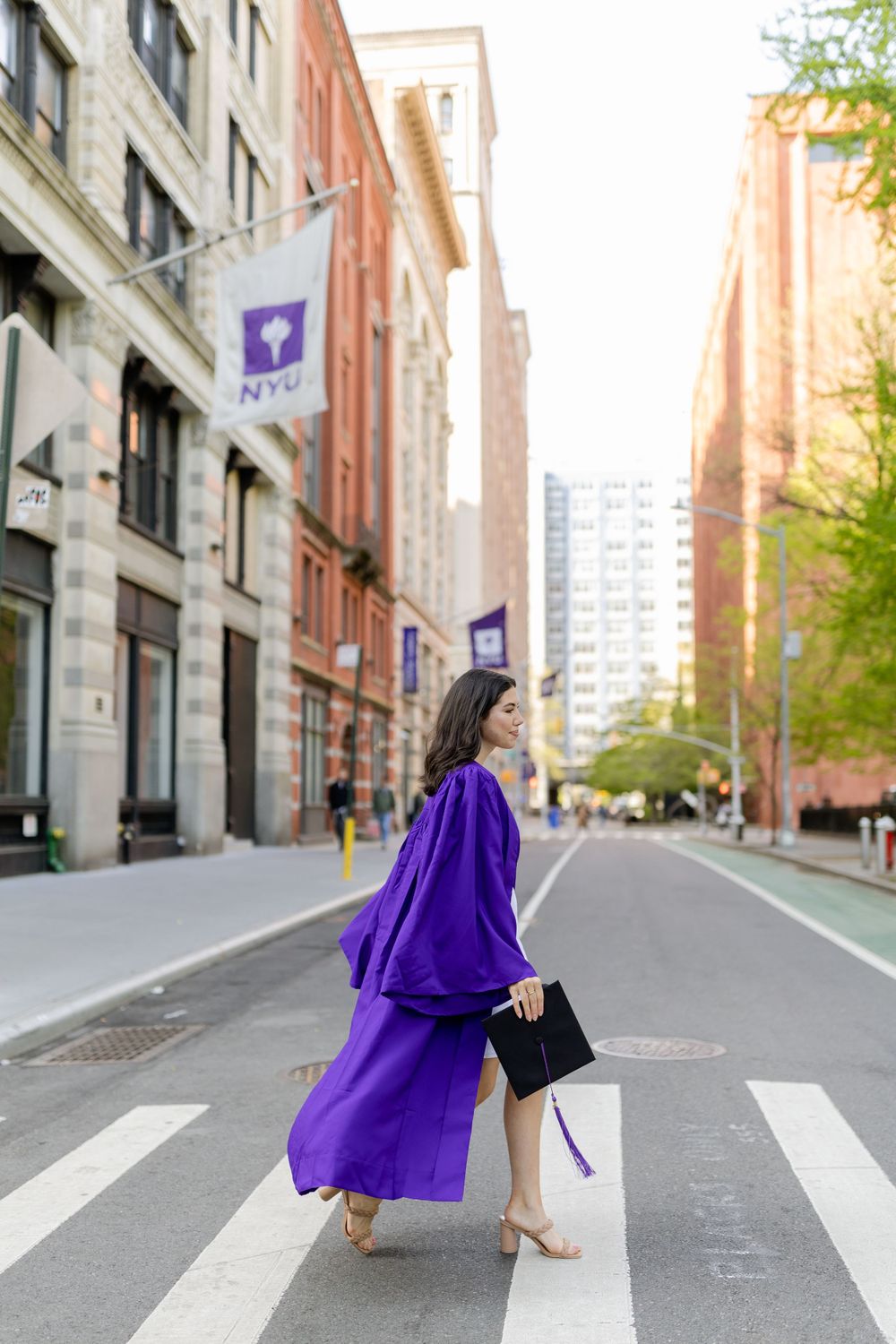 Graduate in purple NYU gown poses on campus street with university flags visible.