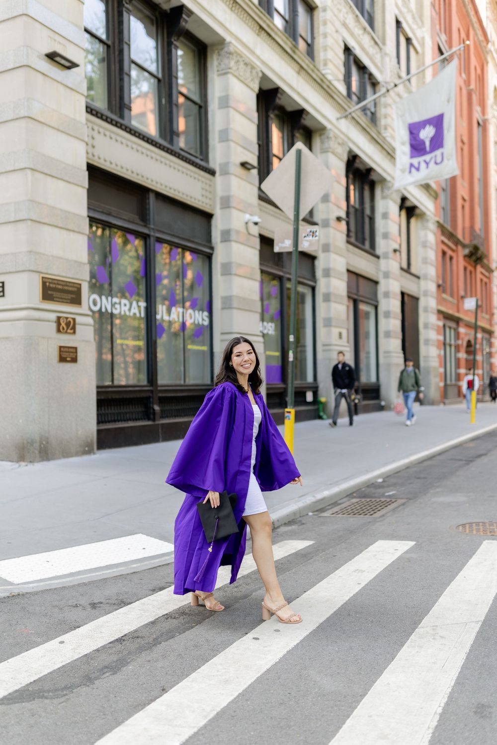Graduate in purple graduation gown crosses street near NYU campus building.