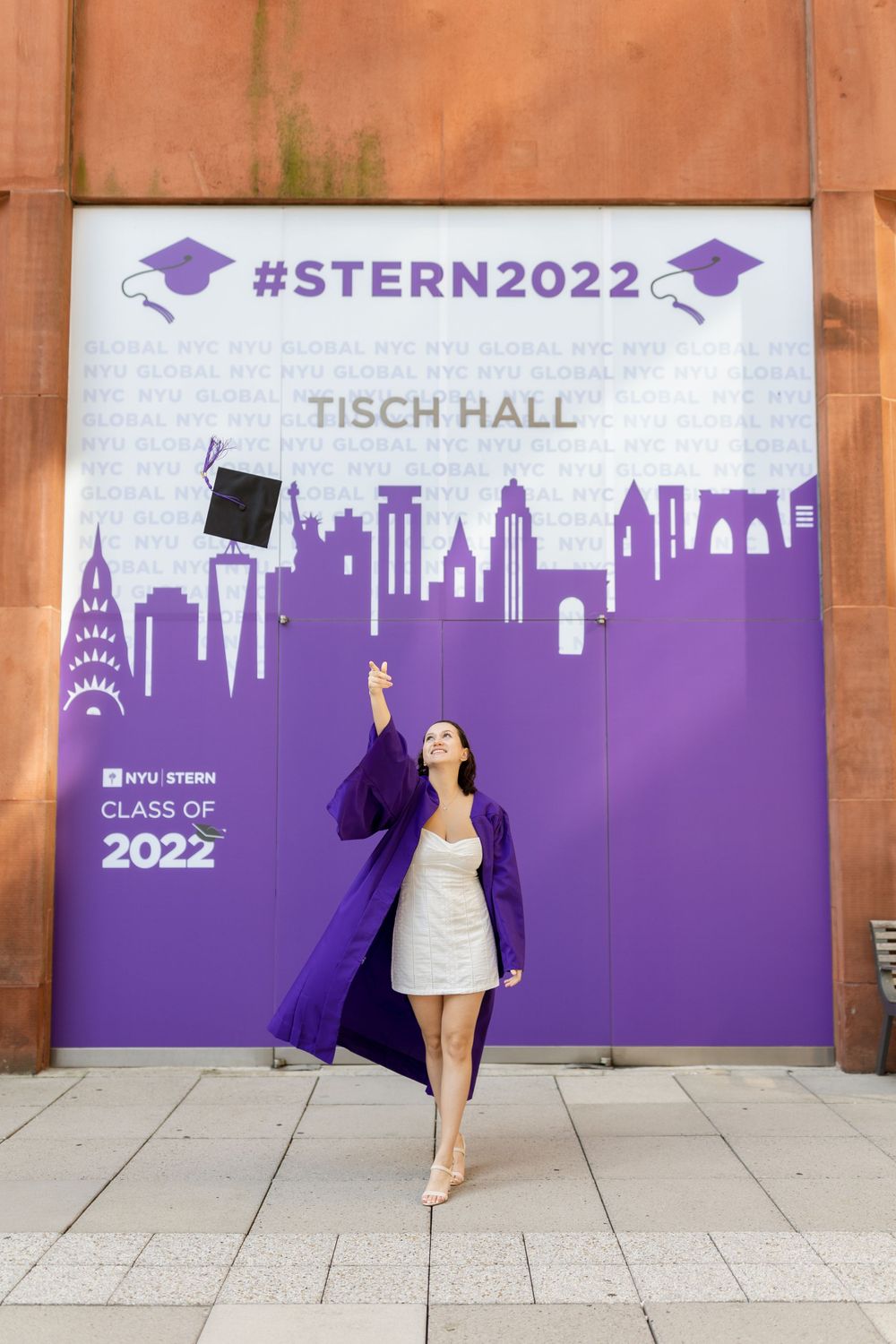A graduate celebrates near Tisch Hall with purple NYU Stern 2022 backdrop showing New York City skyline.