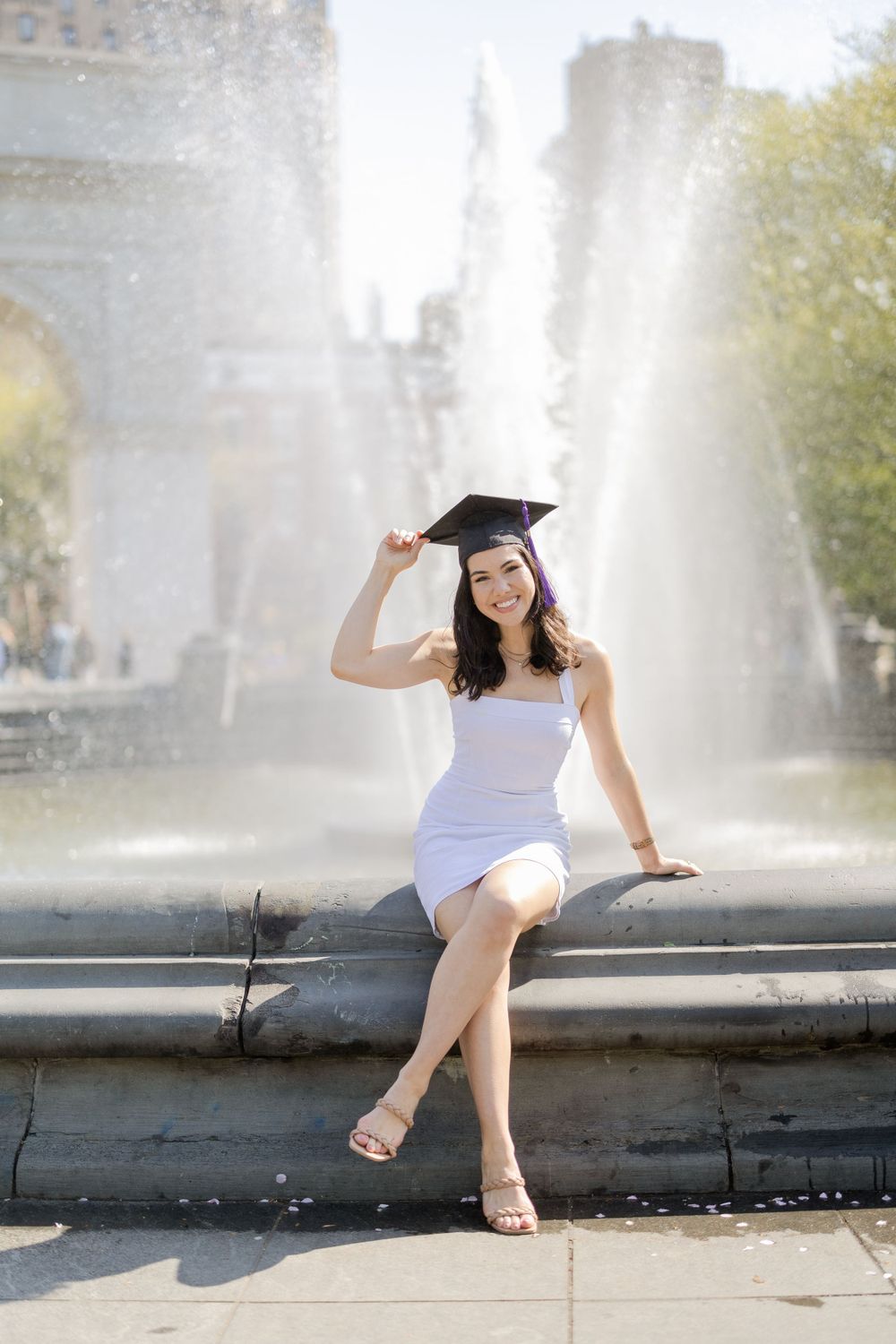 Graduate in white dress sitting by Washington Square fountain.