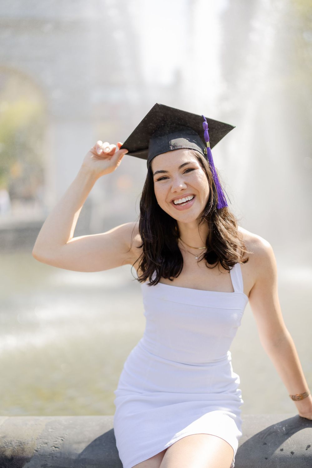 Graduate in white dress and cap poses by fountain on sunny day.