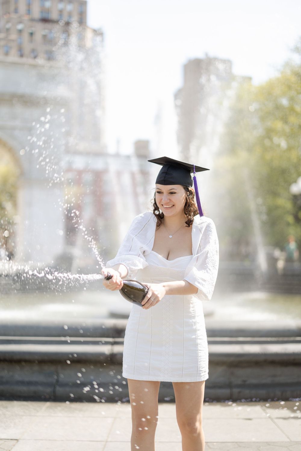 Graduate in white dress popping champagne by fountain.