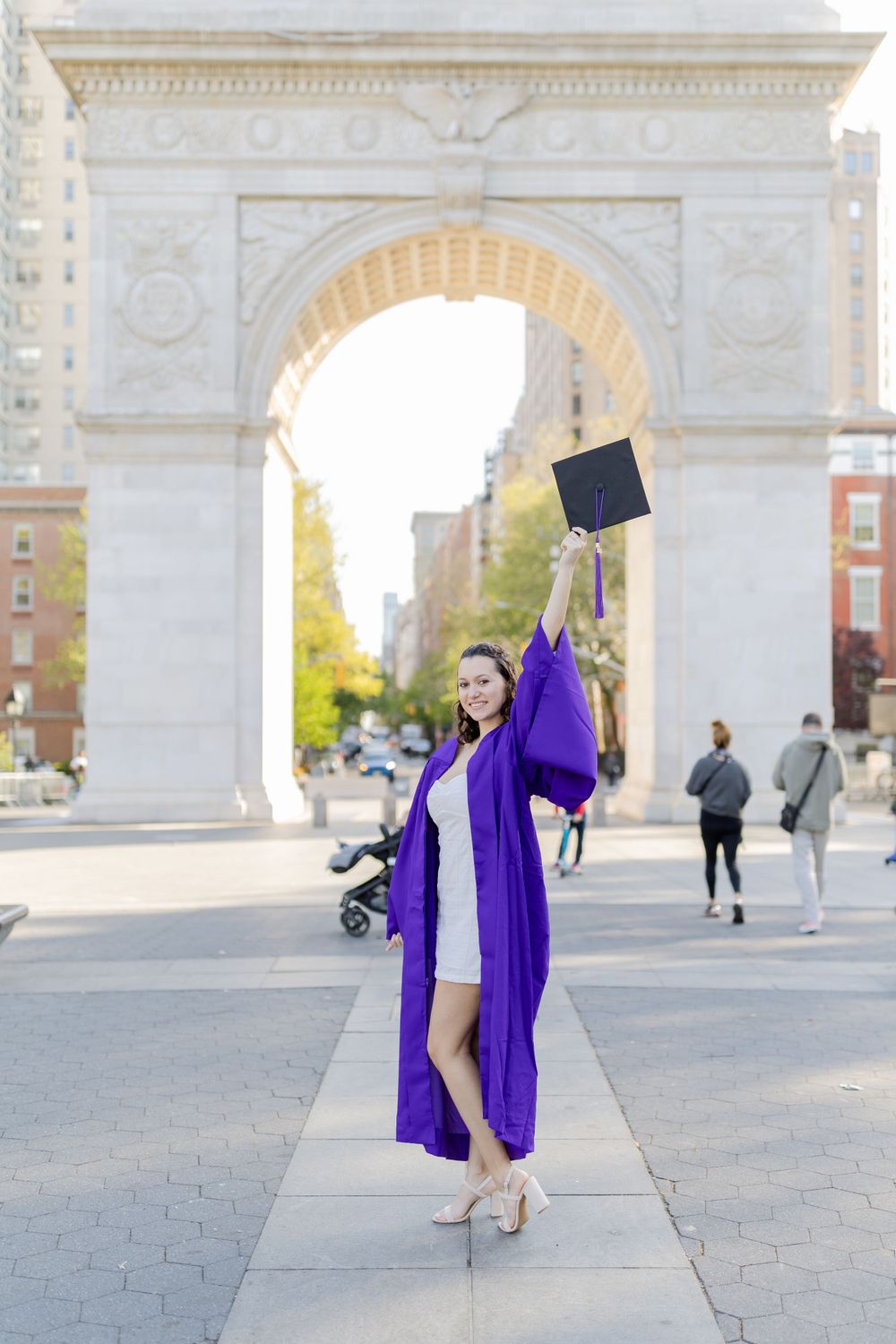 Graduate in purple gown raises cap triumphantly in front of Washington Square Arch.