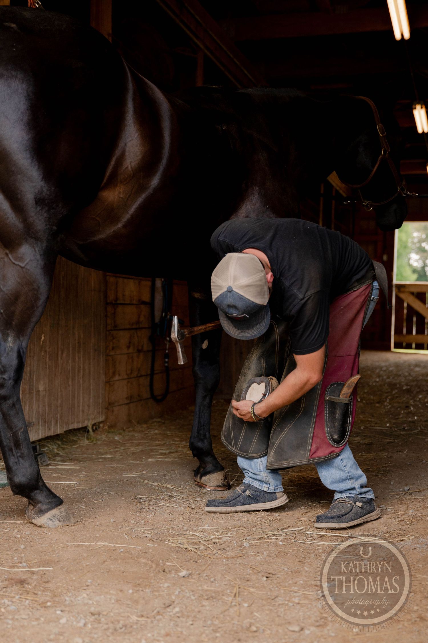 Barn interior showing a farrier performing detailed hoof care on a horse.