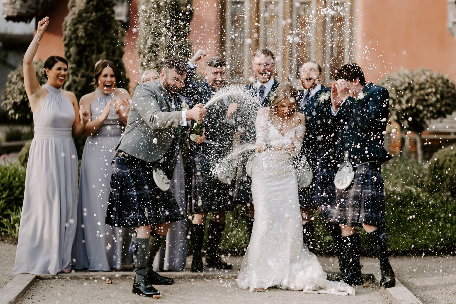 Wedding guests shower the newlyweds with confetti during an outdoor celebration at a venue with peach-colored walls.
