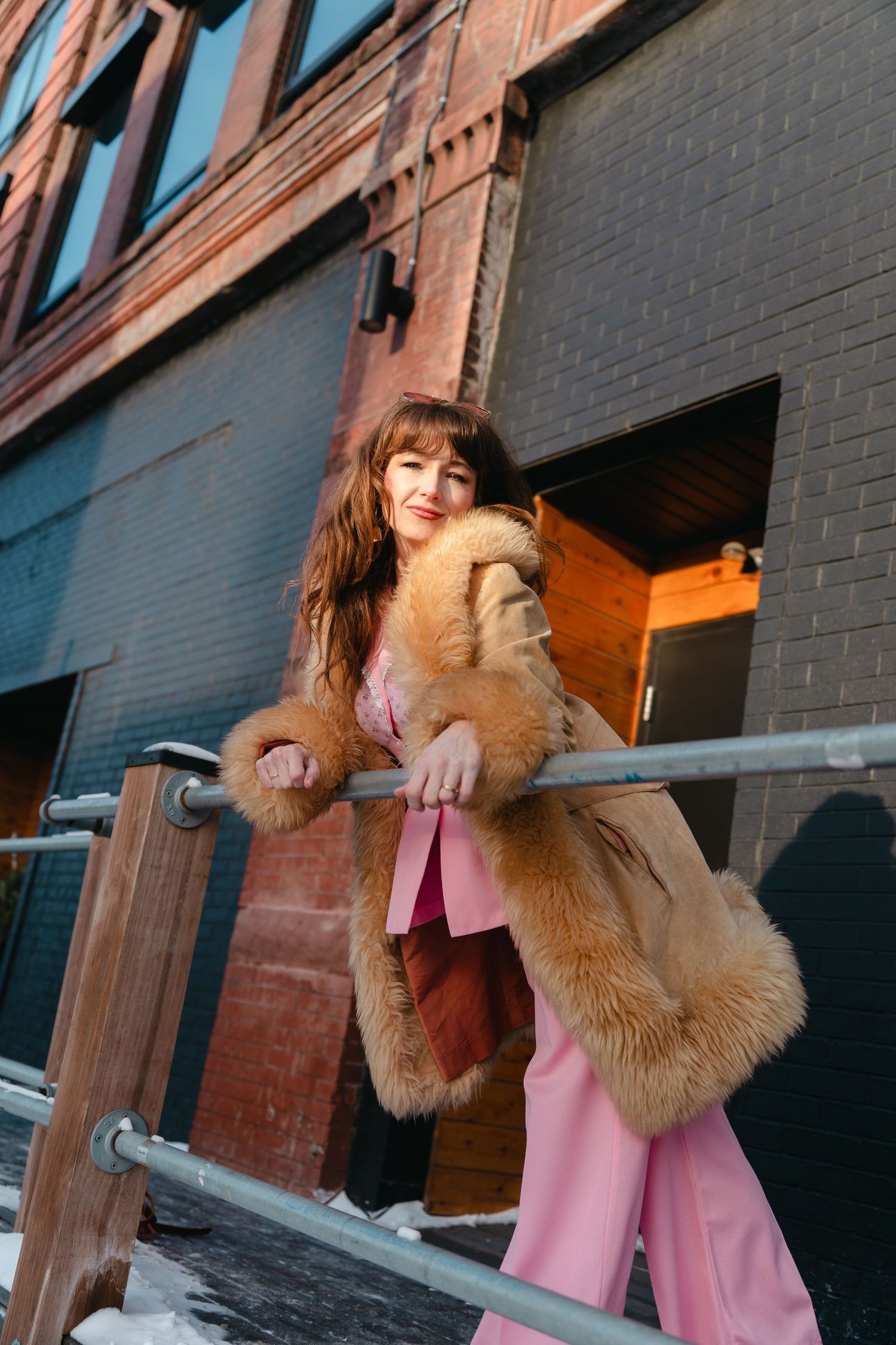 Person wearing a brown fur coat and pink pants leans against railing outside urban building in winter setting.