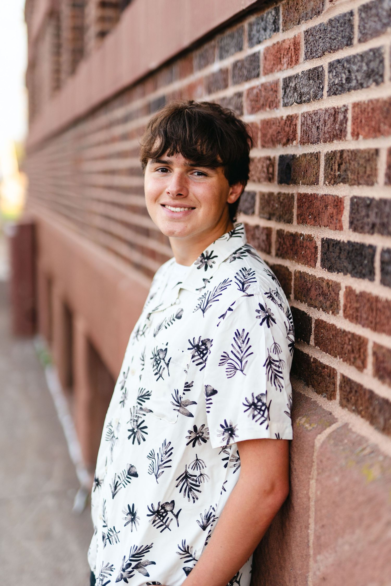 A person wearing a white floral print shirt leans against a brick wall on a sunny day.