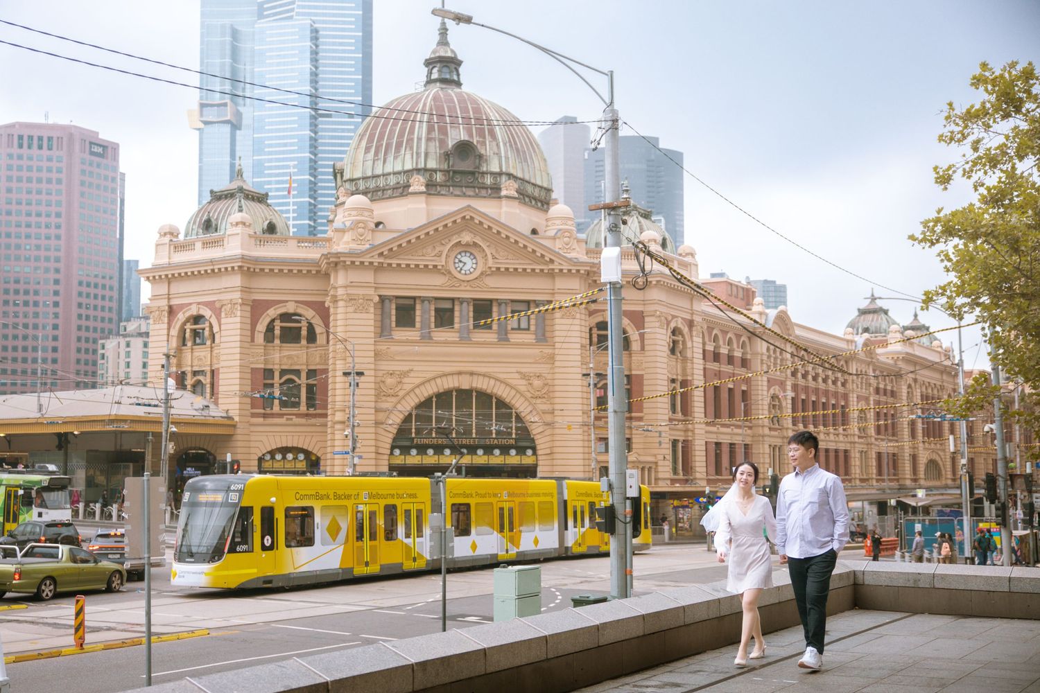 Two people walking past the iconic Flinders Street Station in Melbourne with a yellow tram visible.