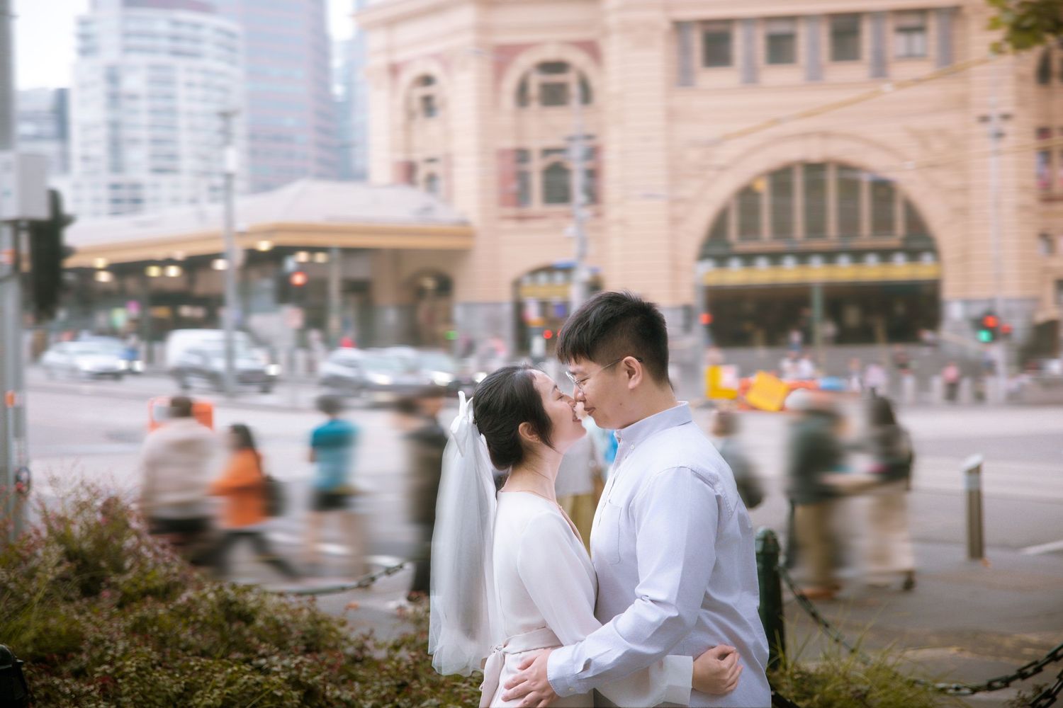 A romantic embrace in front of the historic Flinders Street Station with blurred city background.