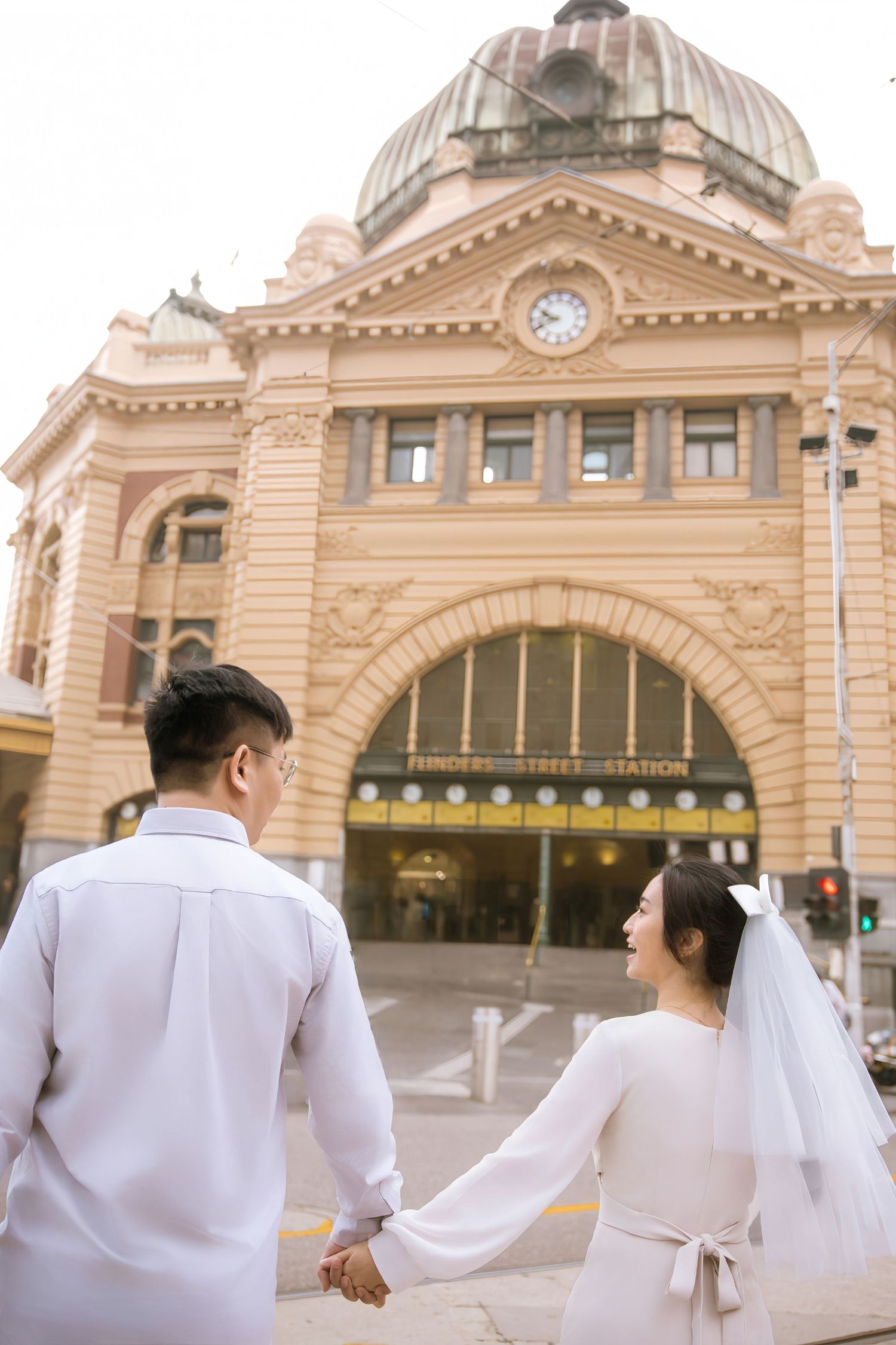 A couple in white attire holding hands in front of Flinders Street Station's iconic dome and clock tower.