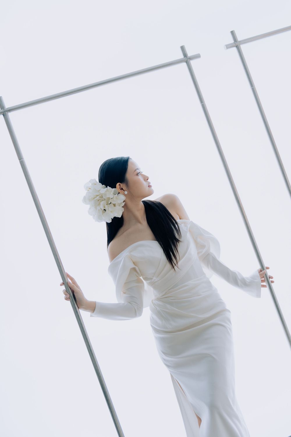 A bride in a sleek white wedding dress and floral hair accessory poses gracefully against a minimalist backdrop.
