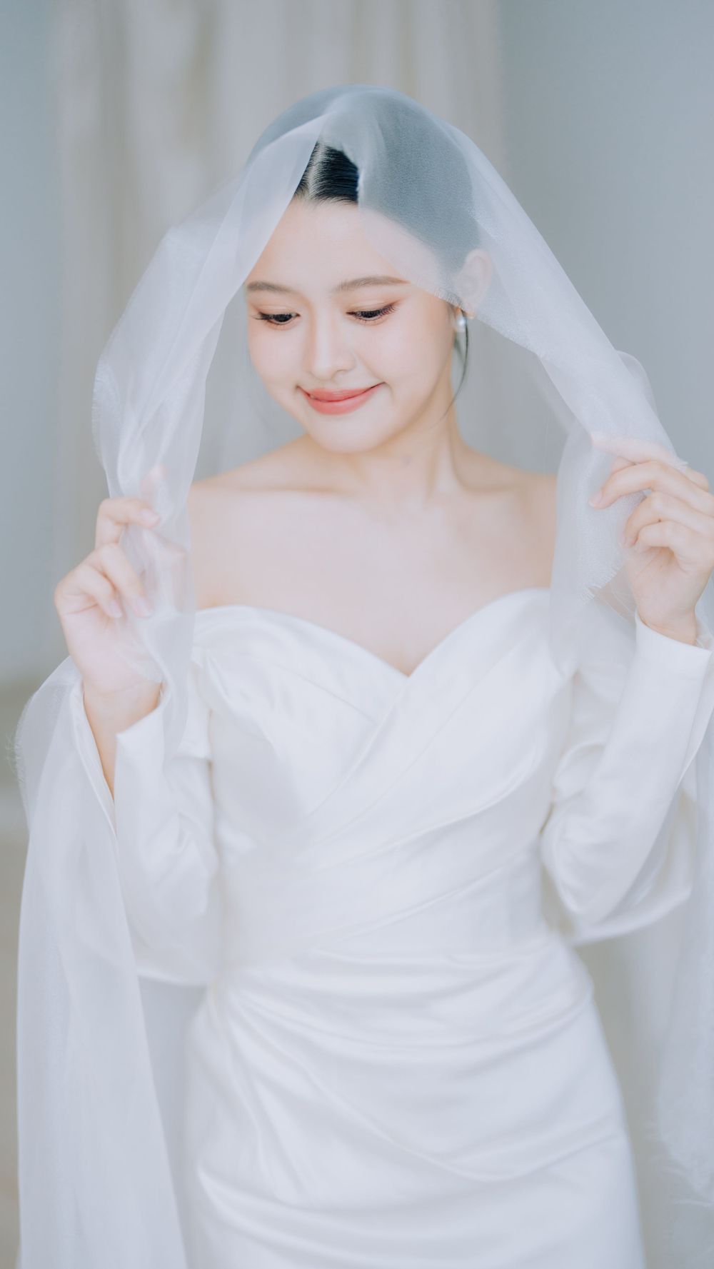 Elegant bride in a white strapless wedding gown gently lifts her delicate veil while smiling softly.