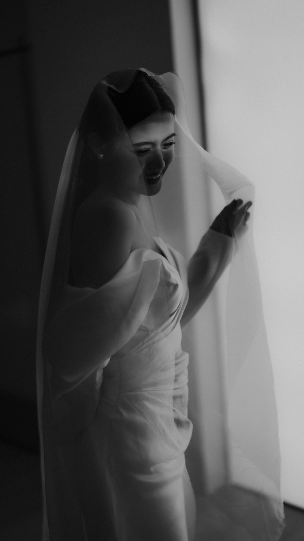 A bride in an elegant white dress stands silhouetted against a window with flowing veil in a black and white photo.