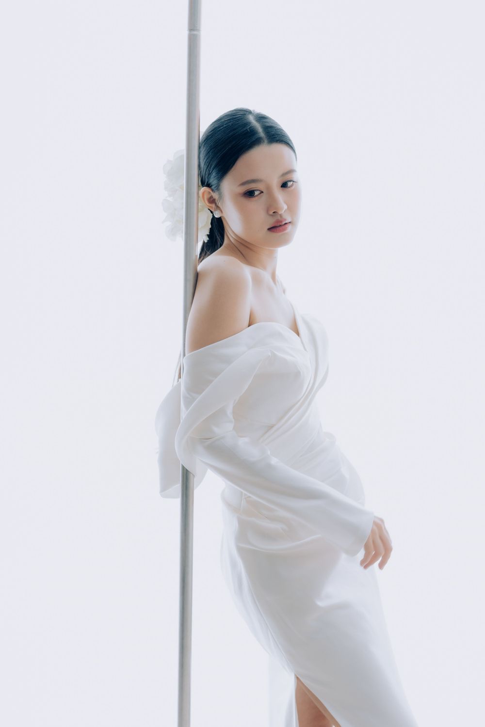 Model in flowing white sheer dress poses gracefully against a minimalist white background with a pole.