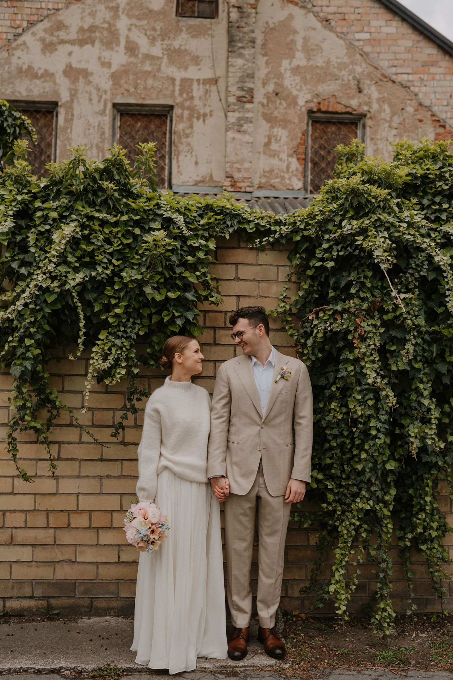 Wedding couple poses together against a rustic brick wall with climbing vines.