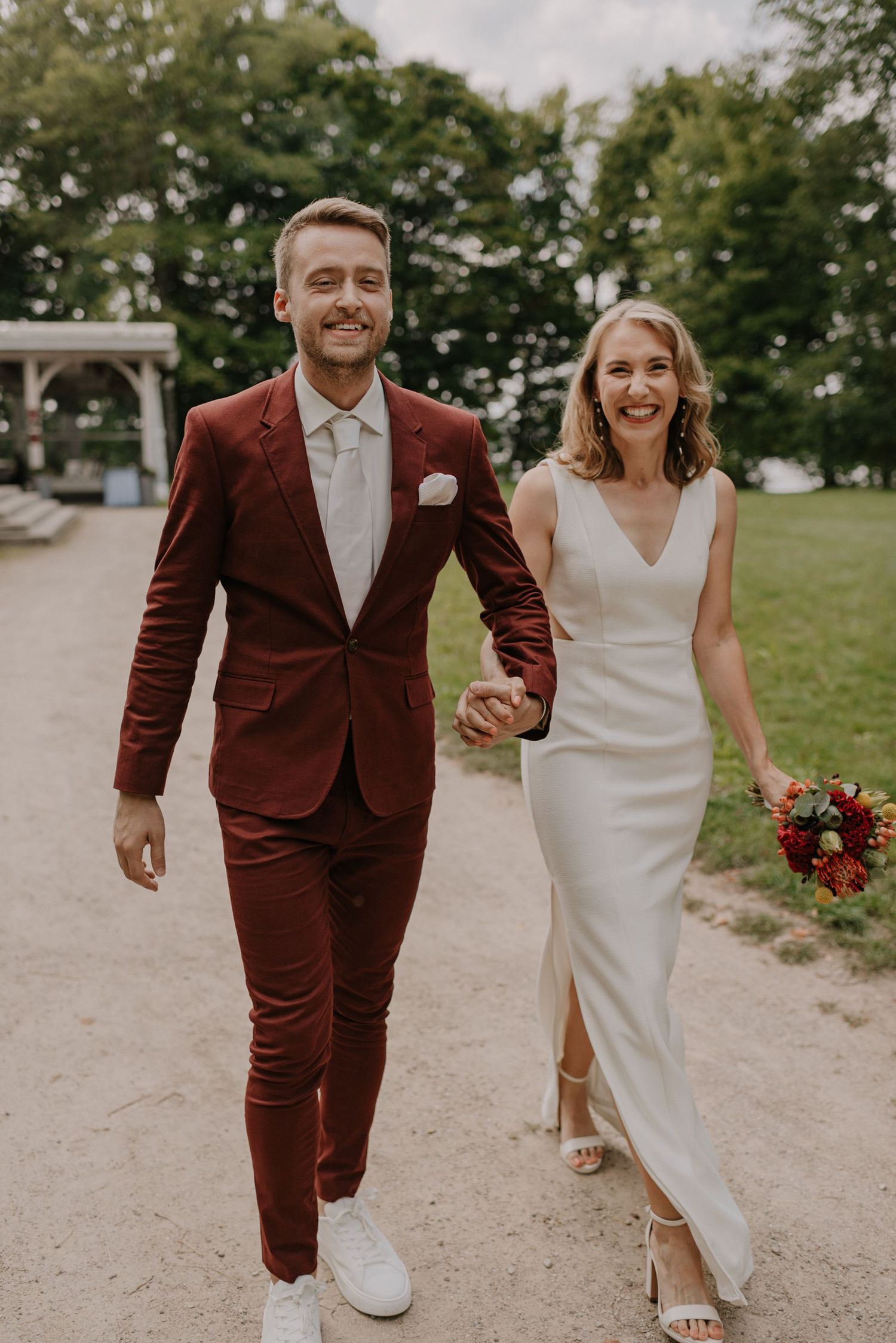 Newlyweds walking together, groom in burgundy suit and bride in elegant white dress.